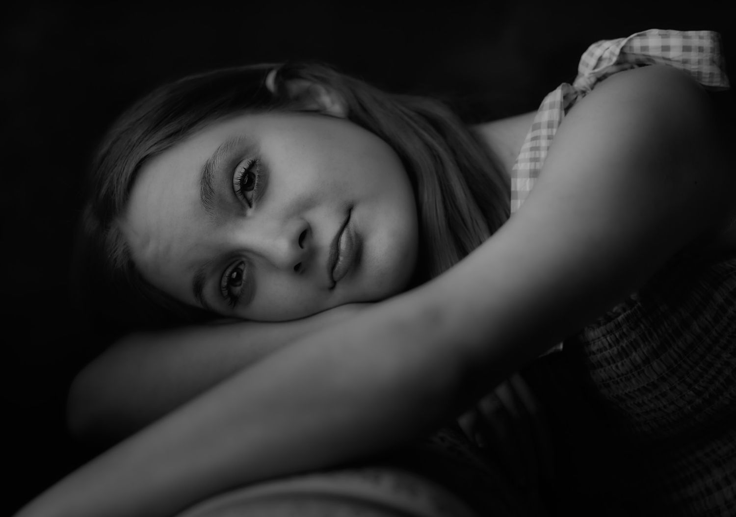 A black and white portrait of a young girl resting her head on her folded arms and gazing softly at the camera.