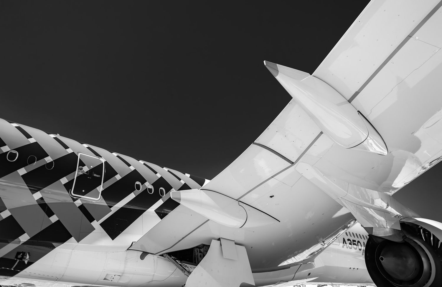 Black and white low-angle photo of an A350 wing, winglet and jet engine beside a patterned fuselage against a dark sky.