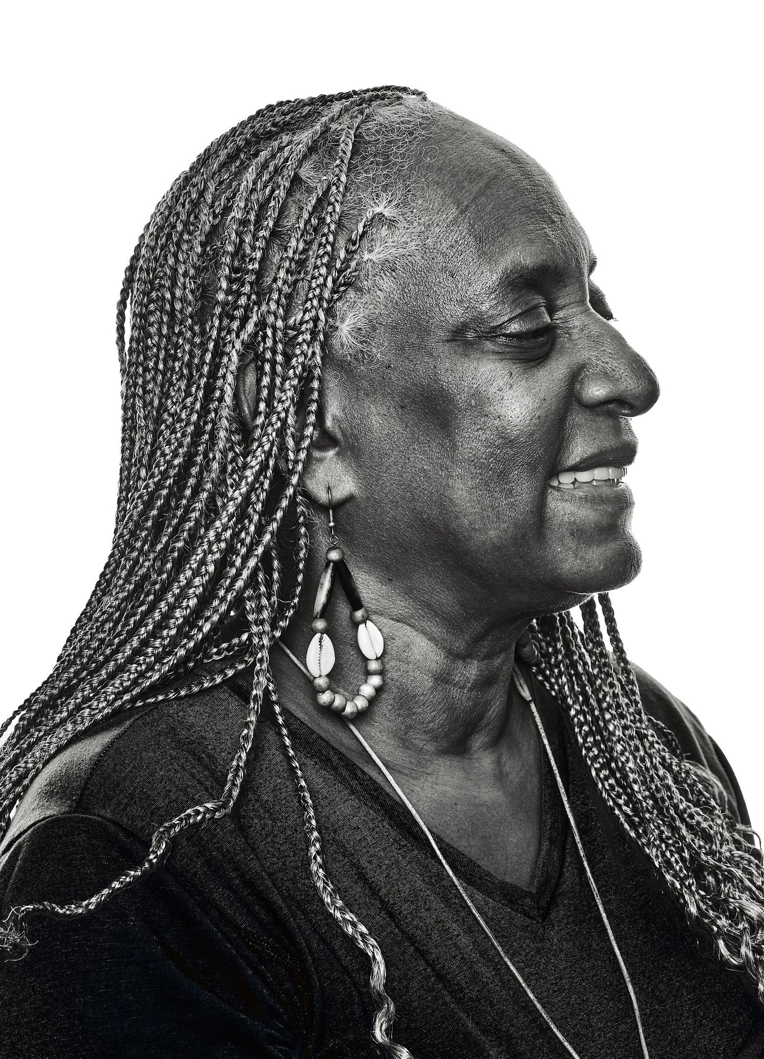 Black and white profile portrait of a smiling older woman with long braids, drop earrings, and a layered necklace against a white backdrop.