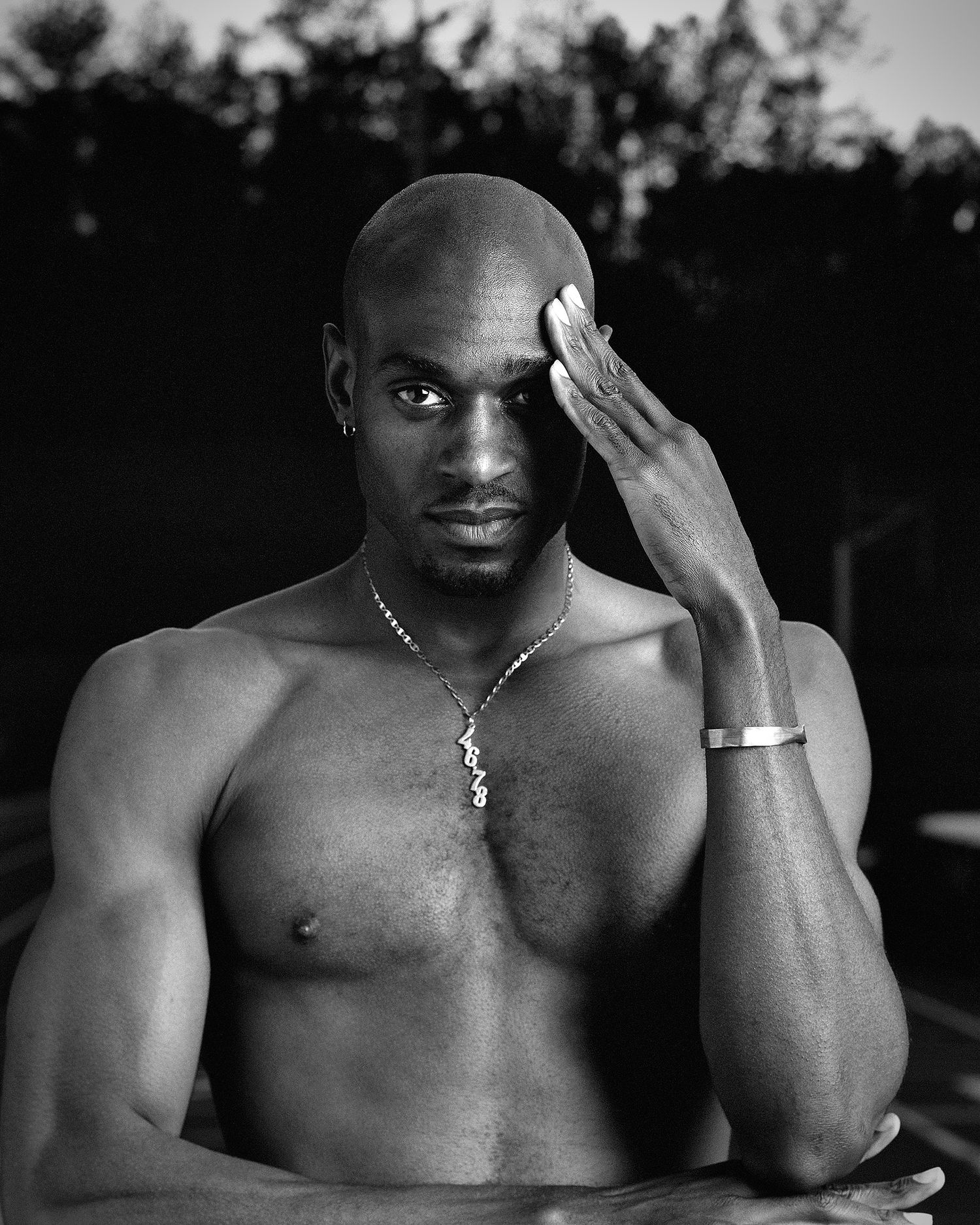 Black and white portrait of a shirtless, muscular bald man wearing a pendant necklace and bracelet, hand raised to his forehead.
