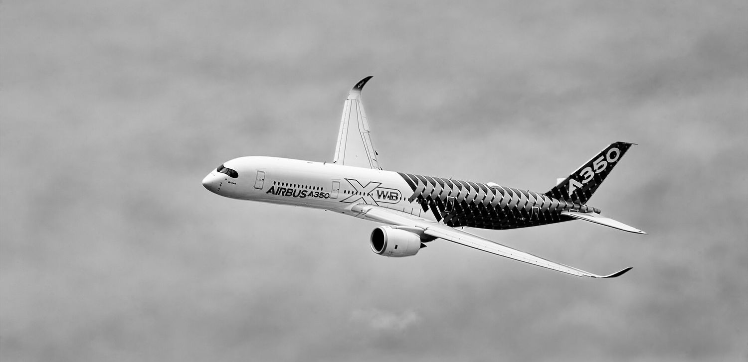 Airbus A350 passenger jet banking in flight against a cloudy sky, showing its carbon fiber fuselage and winglets in black and white.