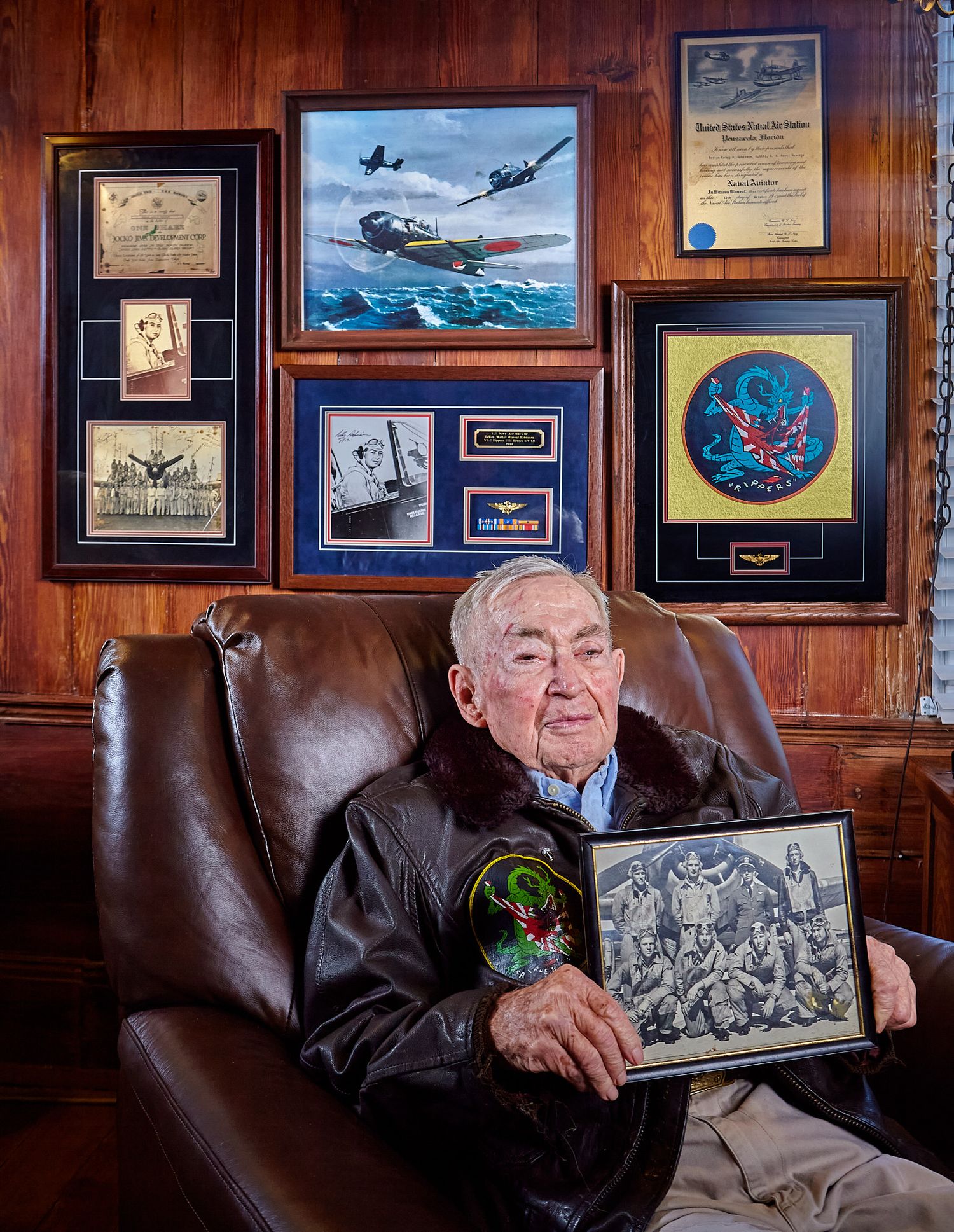 Elderly veteran in a leather bomber jacket sits in a brown armchair holding a framed black-and-white crew photo beside aviation memorabilia.