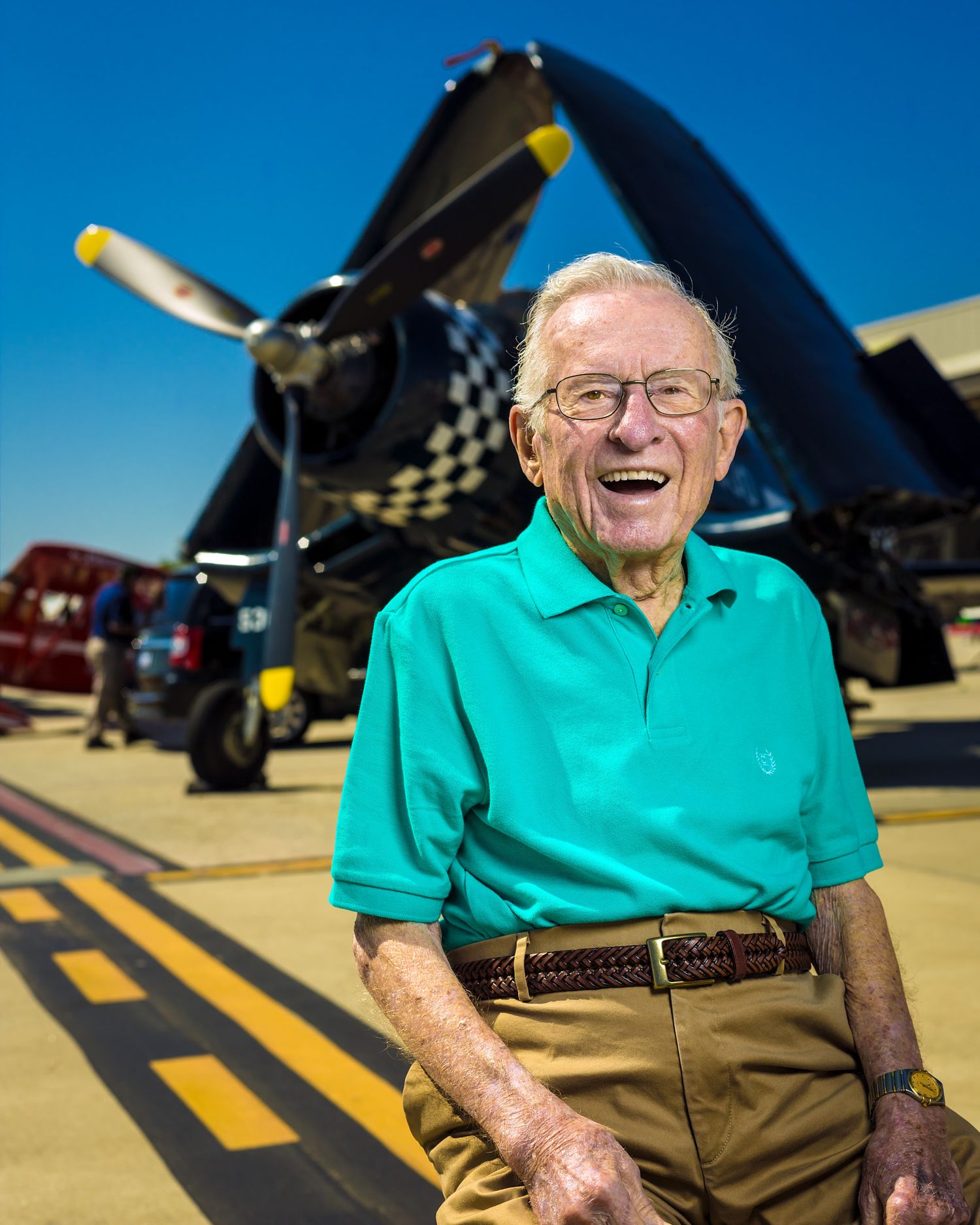 Smiling elderly man in a teal polo and khaki pants sits on an airfield in front of a vintage Corsair warplane with checkered cowling.