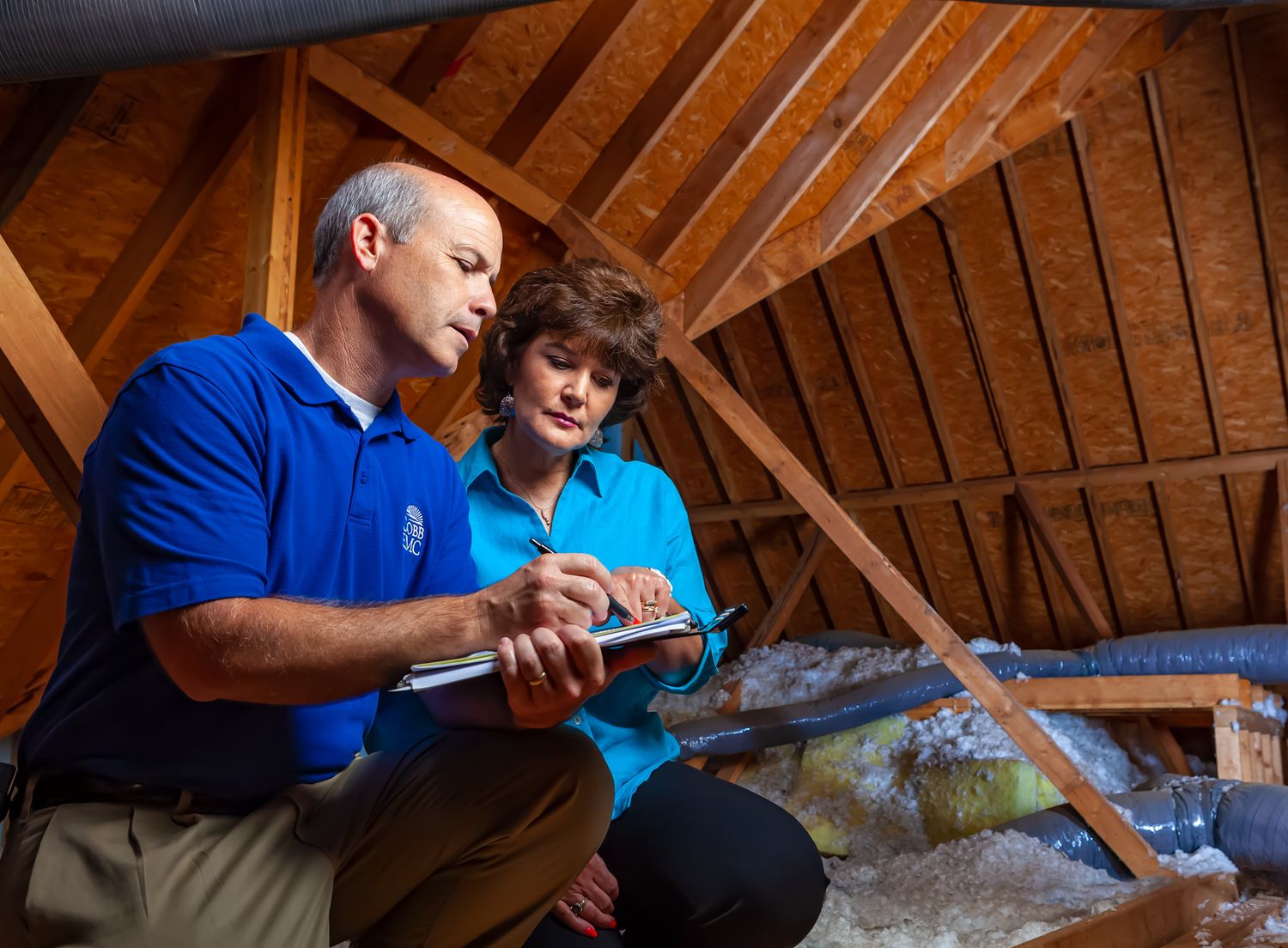 Two home inspectors in blue shirts review a clipboard in an attic with exposed roof rafters and insulation.
