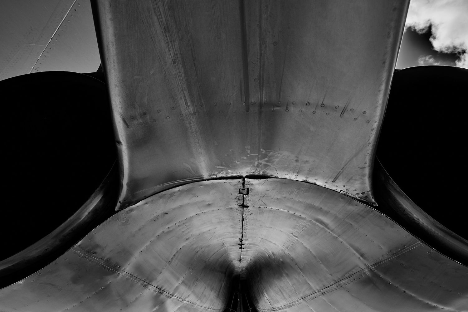 Black and white view of a riveted metal aircraft engine pylon and nacelle seen from below against a dramatic sky.