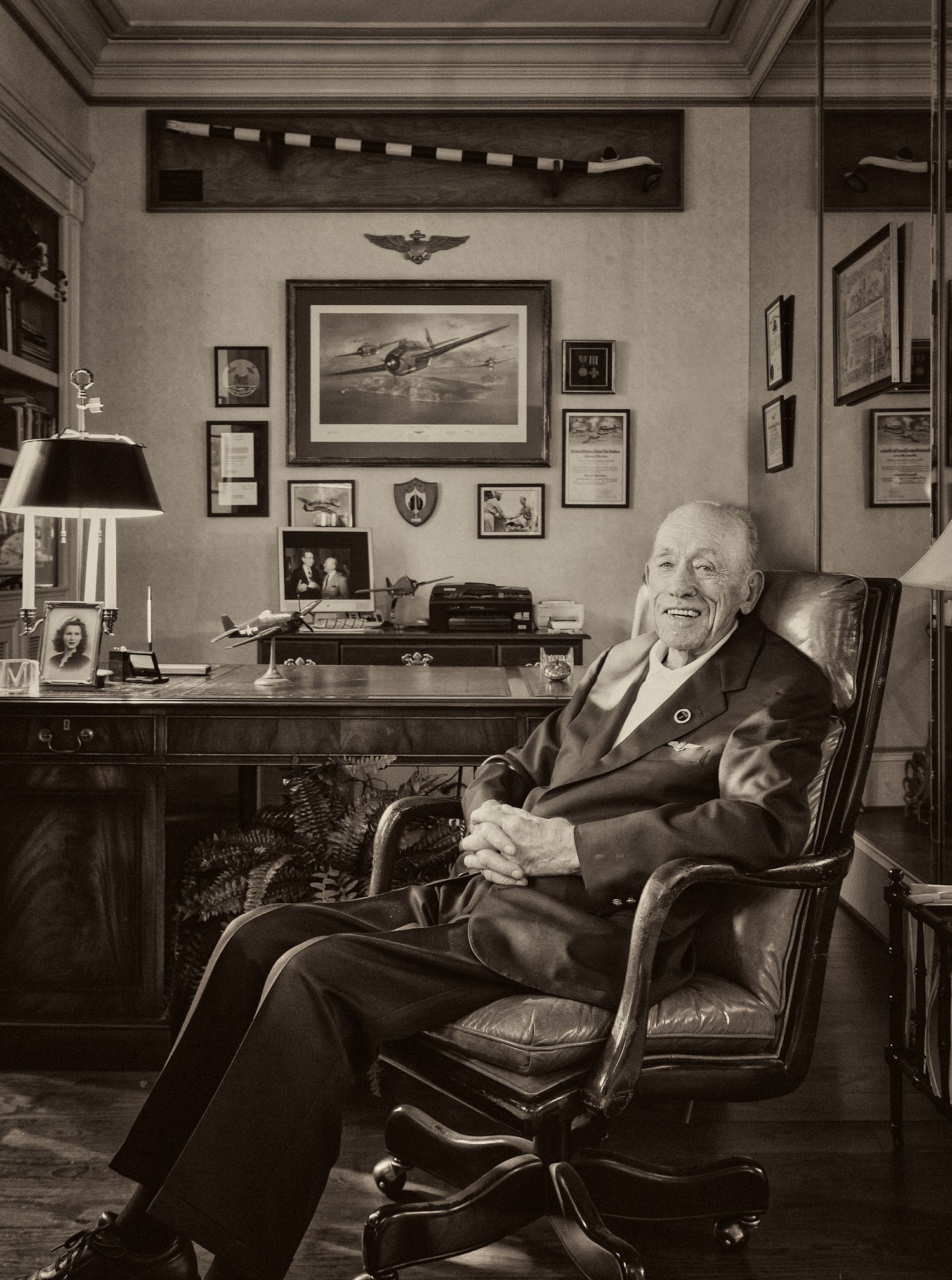 Black and white portrait of a smiling elderly man in a suit seated at a wooden desk in an office filled with aviation memorabilia.