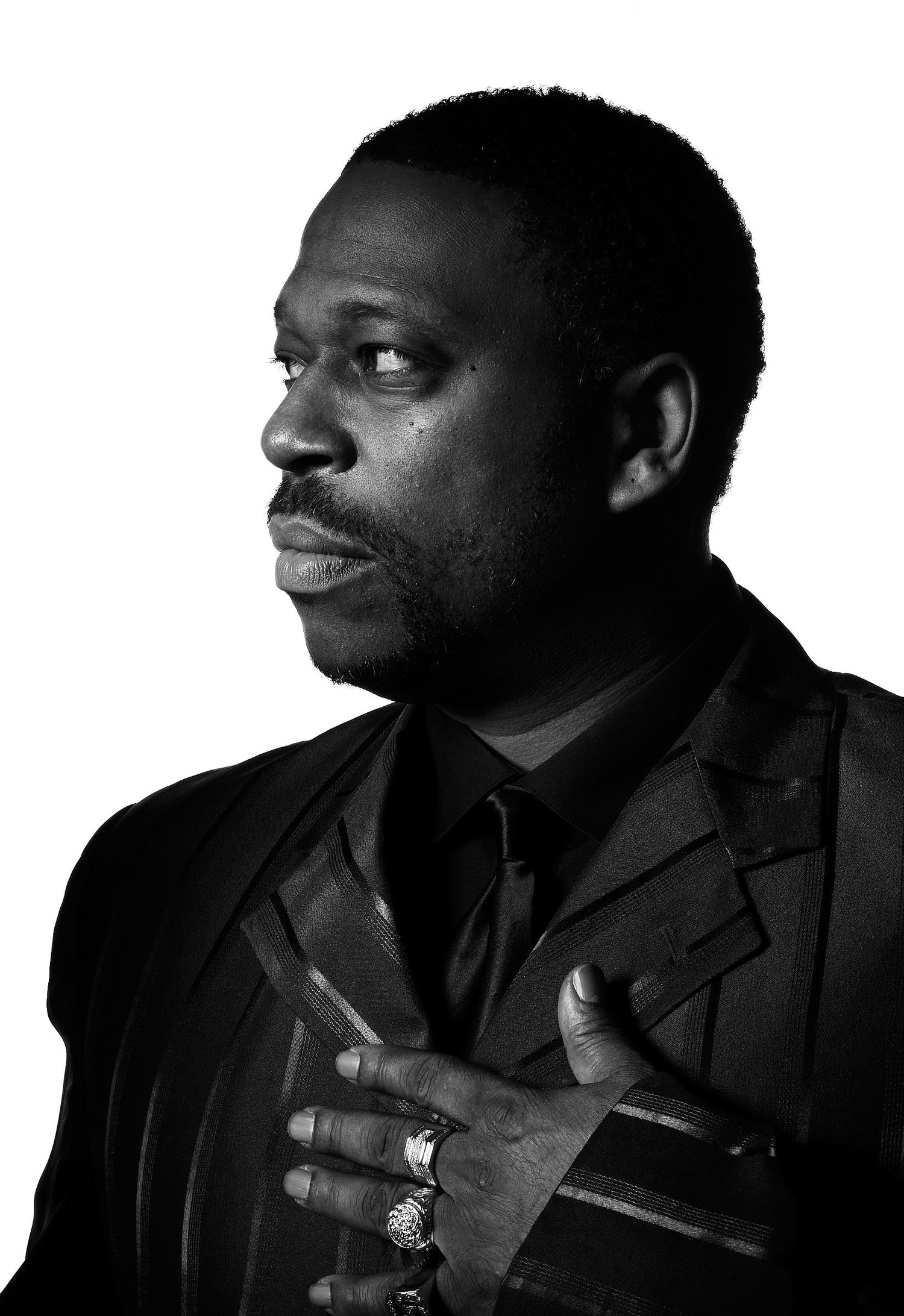 Black and white portrait of a man in a dark pinstriped jacket and tie, gazing to the side with his hand over his chest, wearing rings.