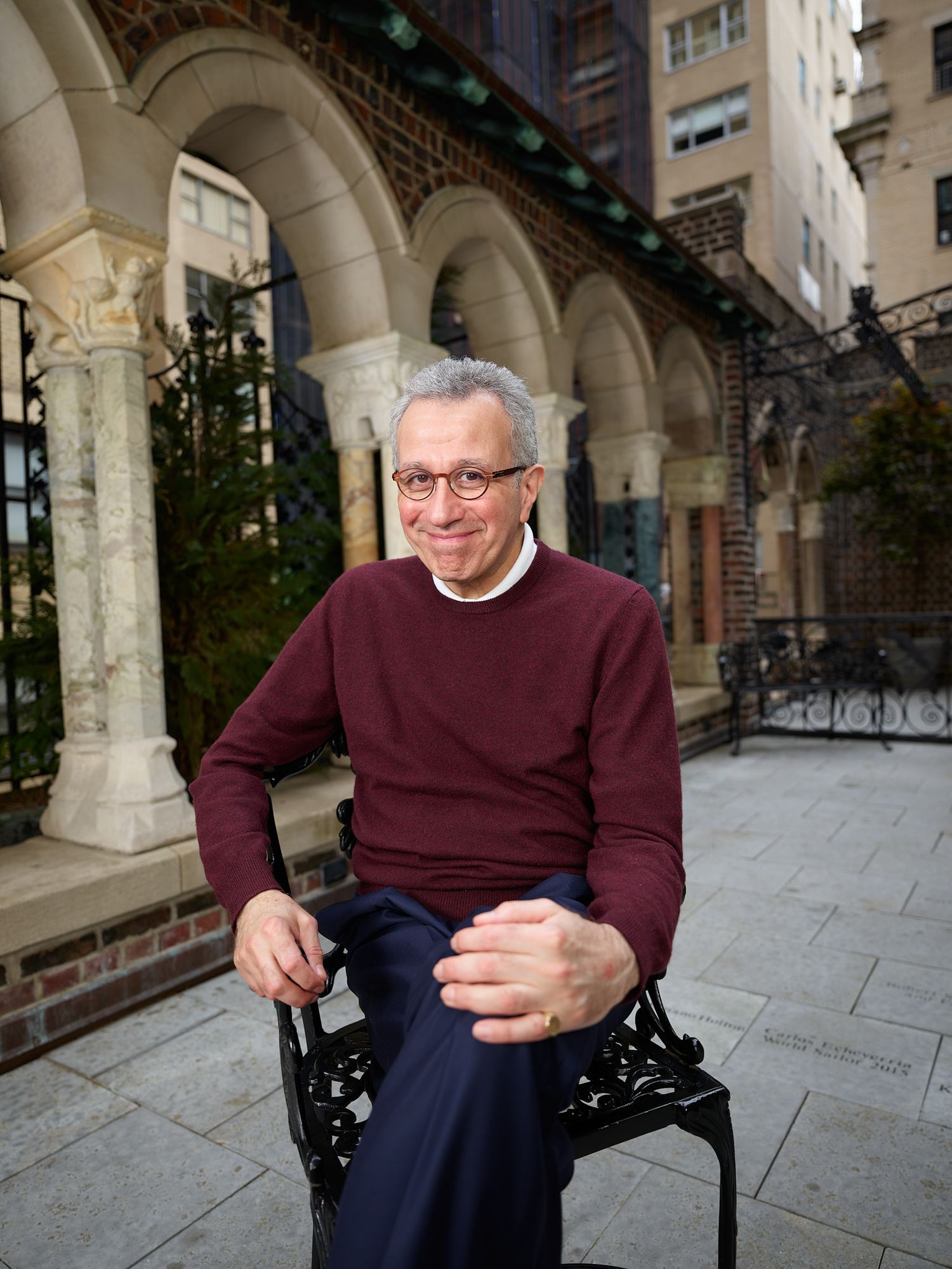 Smiling man with gray hair and glasses in a maroon sweater sits on an ornate black chair in a stone courtyard with arched columns.