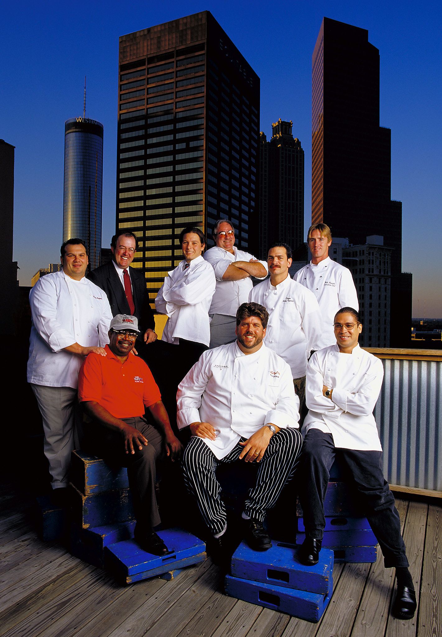 Group of nine chefs in white coats posing on a rooftop terrace at dusk with a glowing city skyline and tall skyscrapers behind them.