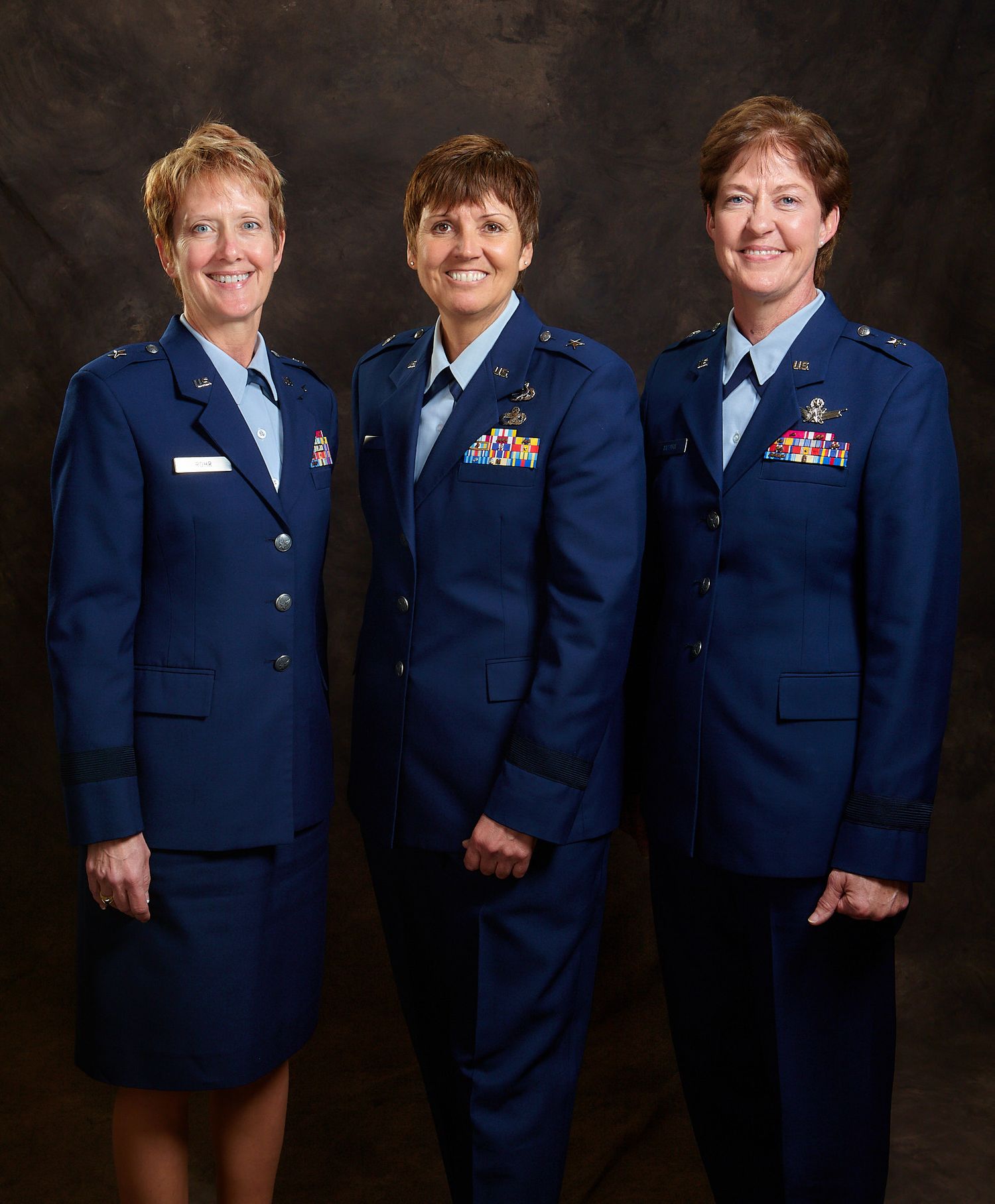 Three smiling women in blue Air Force dress uniforms with ribbons and insignia pose together for a formal portrait against a dark backdrop.
