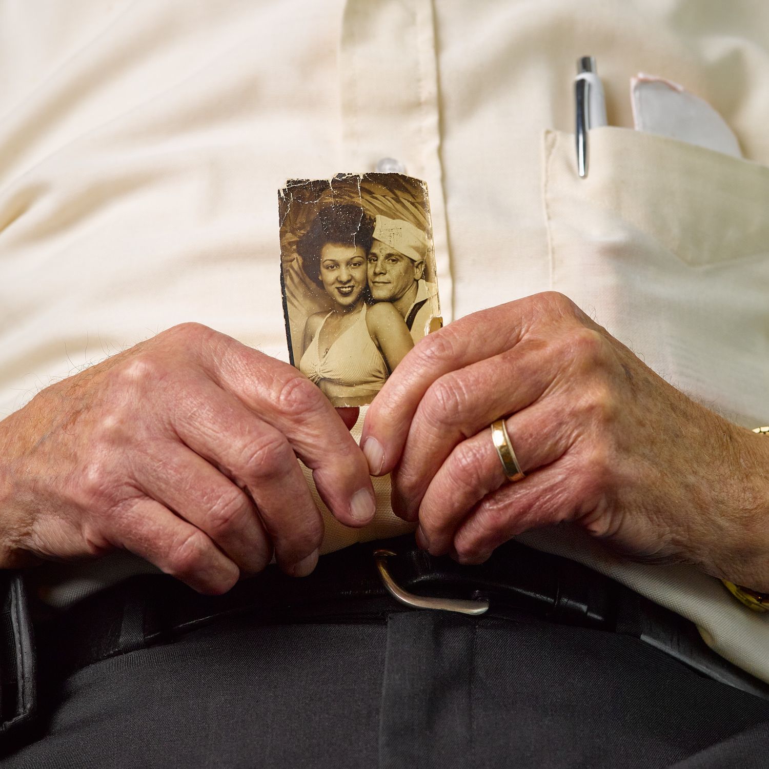 Elderly man's weathered hands holding a vintage sepia photo booth portrait of a smiling woman and a sailor, a pen in his shirt pocket.