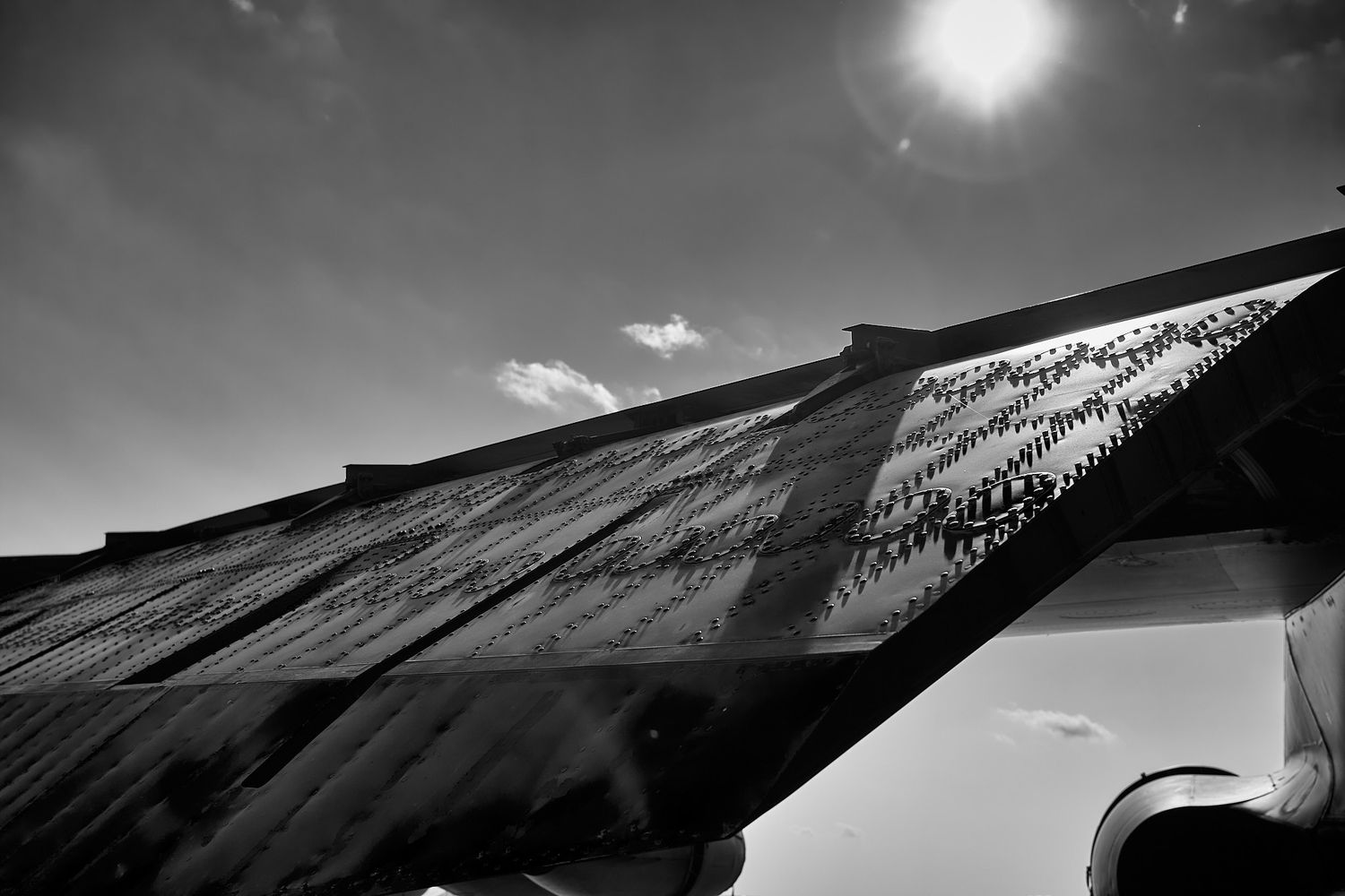 Sunlit aircraft wing with textured surface patterns photographed in black and white against a bright sun and scattered clouds.