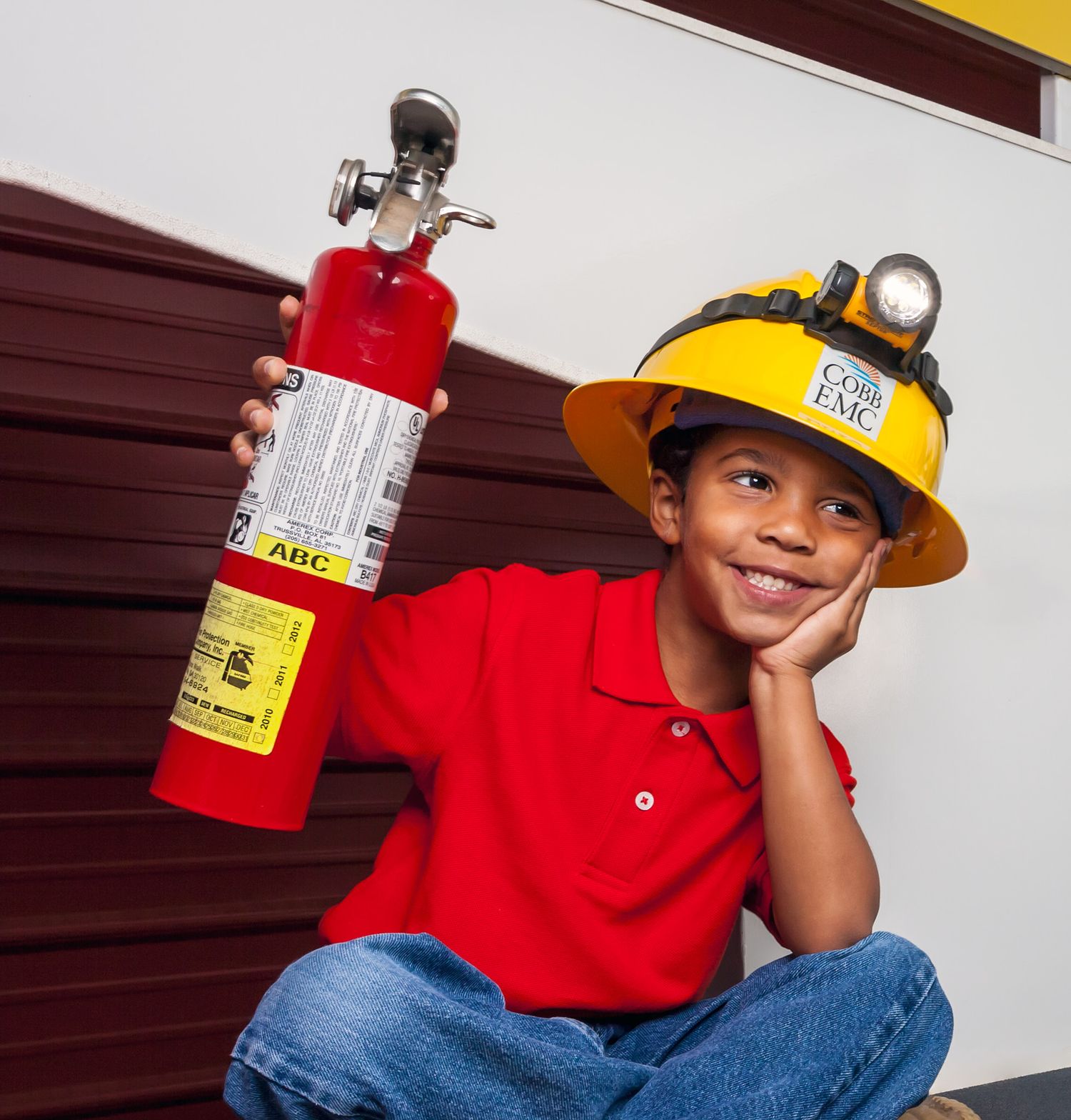 Smiling boy in a yellow EMC hard hat with headlamp and red polo holds a red ABC fire extinguisher while resting his chin on his hand.