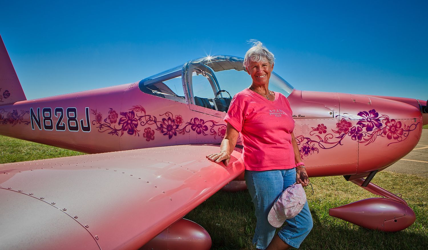A smiling older woman in a pink shirt stands beside a pink experimental airplane with purple flower decals and registration number N828J.