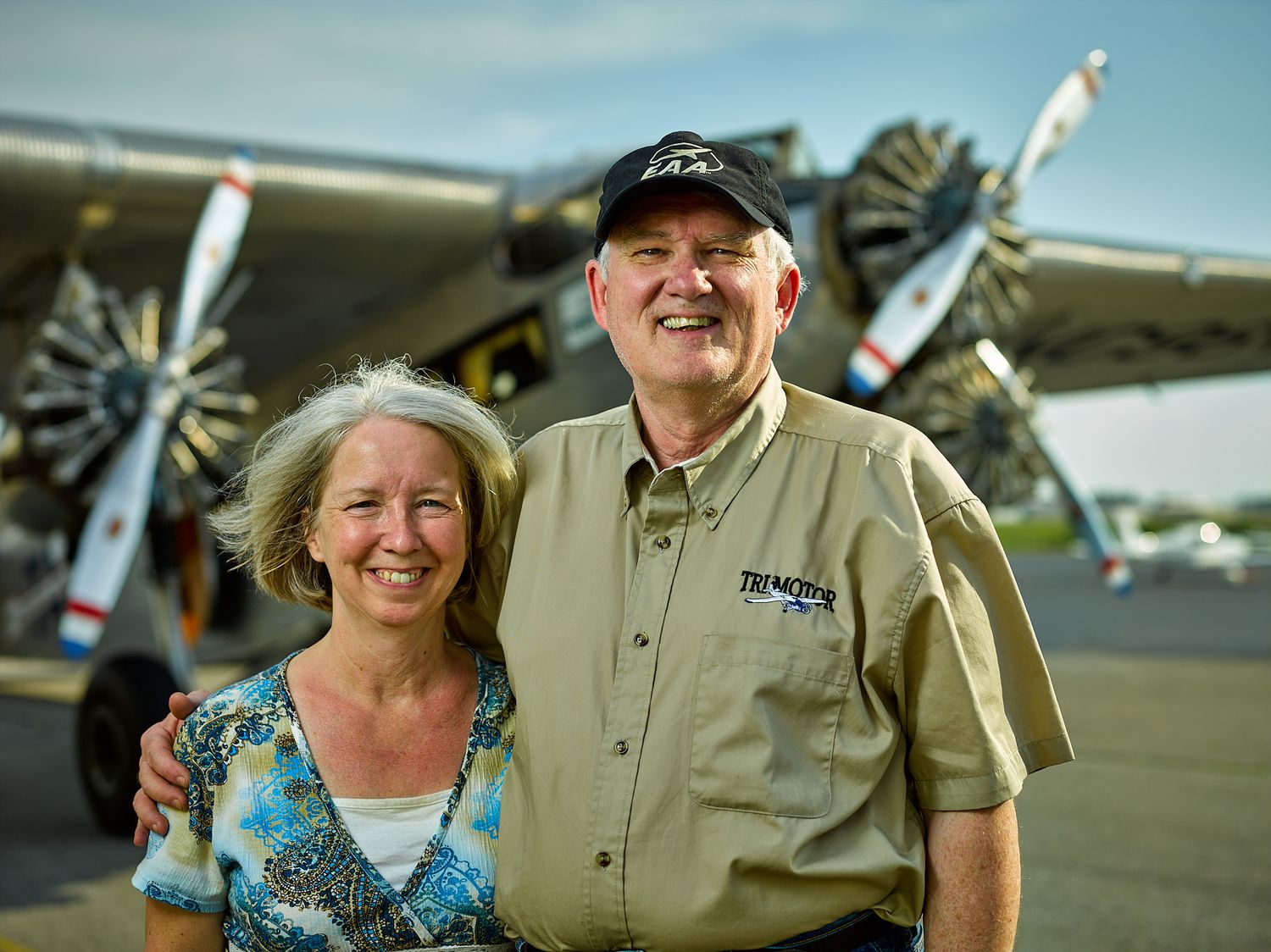 Smiling man in a black cap and khaki shirt stands beside a smiling woman in a floral top in front of a vintage propeller airplane.
