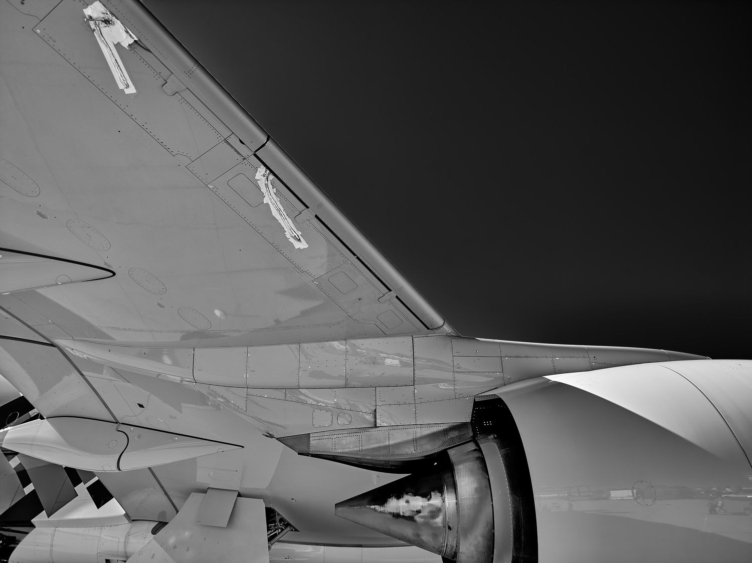 Black and white close-up of an airplane wing and jet engine nacelle with flaps and panel details against a dark sky.