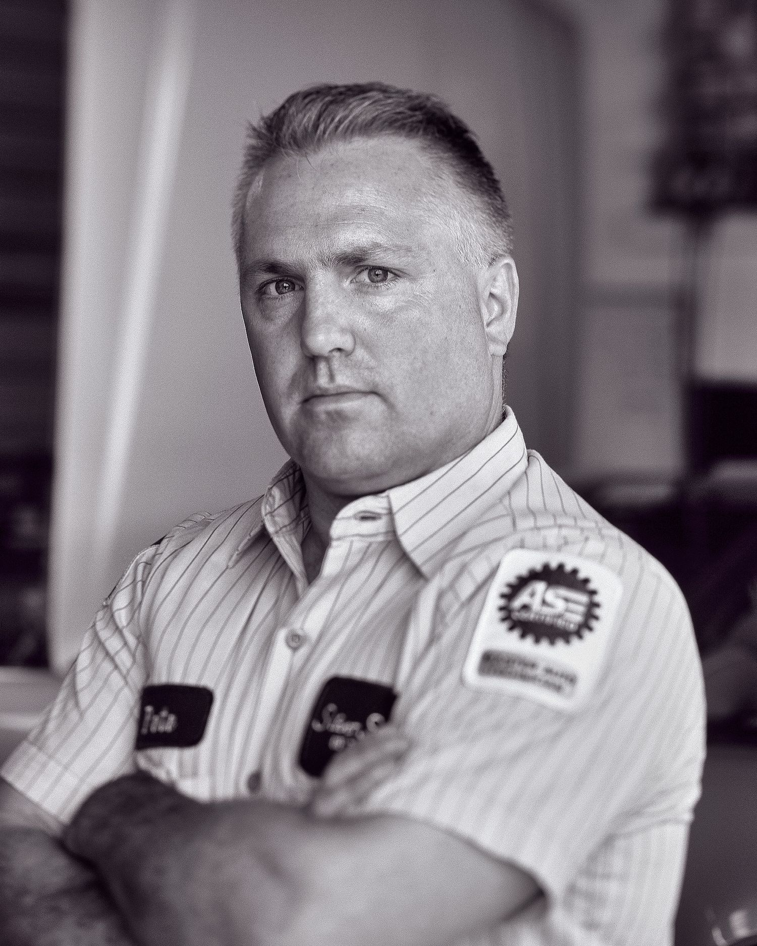 Black and white portrait of a serious mechanic with short hair and crossed arms, wearing a striped work shirt with an ASE patch.