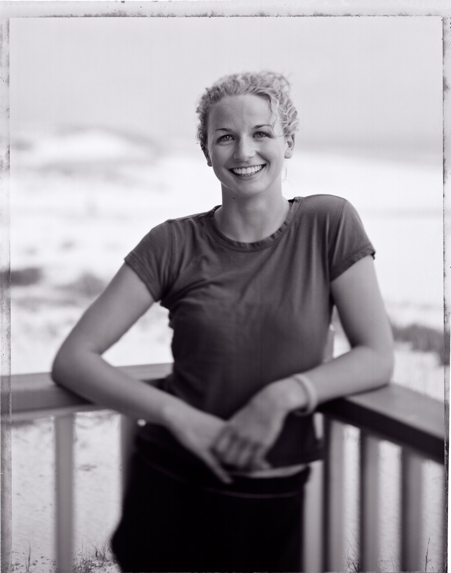 Smiling woman with curly hair leans on a wooden porch railing, with a sandy beach and dunes softly blurred behind her in black and white.