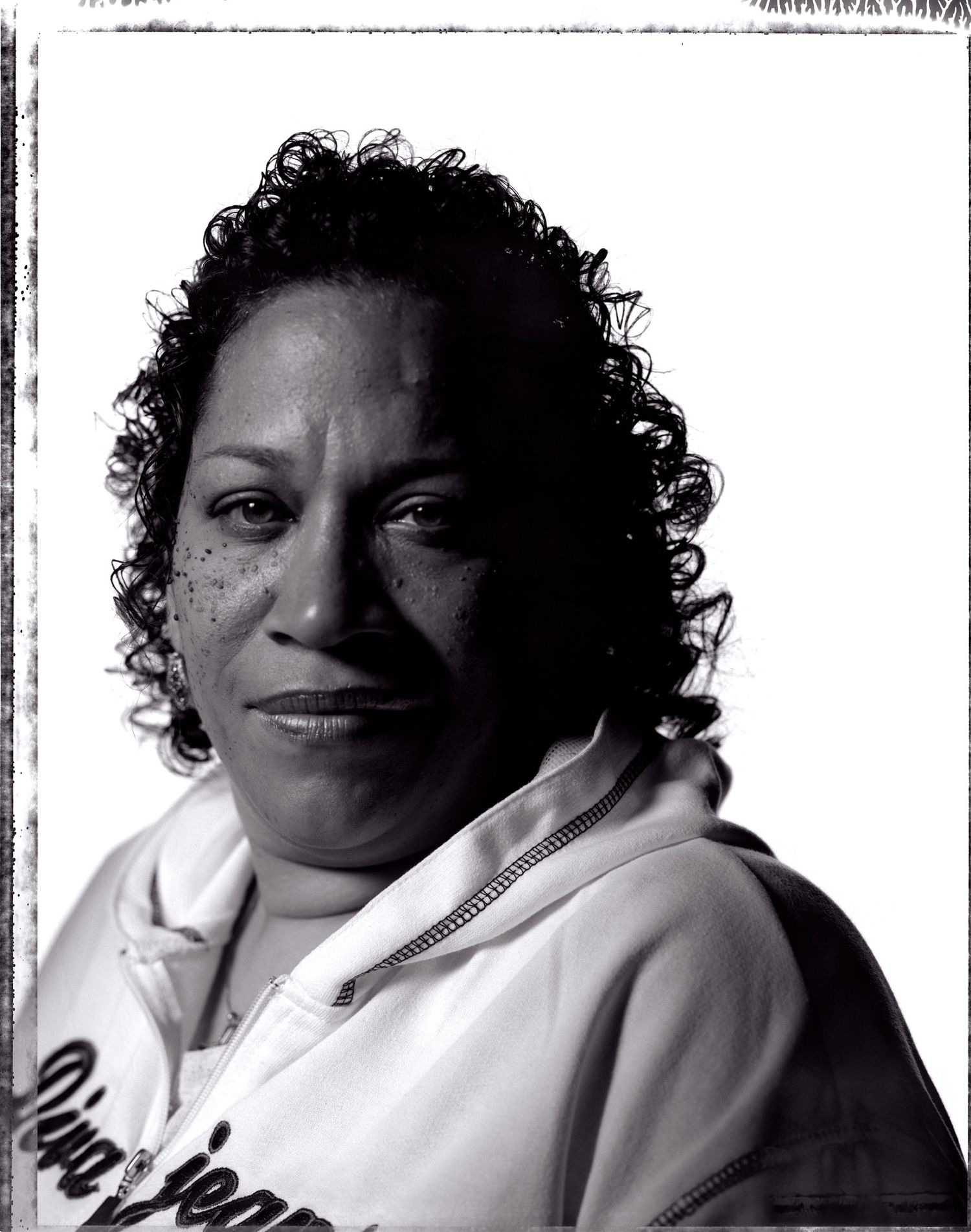 Black and white studio portrait of a woman with short curly hair wearing a white zip hoodie, looking calmly at the camera.