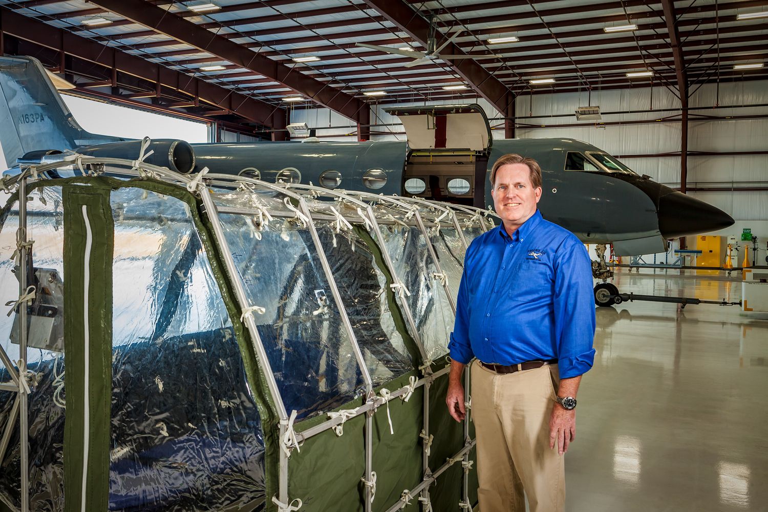 Smiling man in a blue button-up shirt and khakis stands in an aircraft hangar beside a military jet and a clear-paneled canvas shelter.