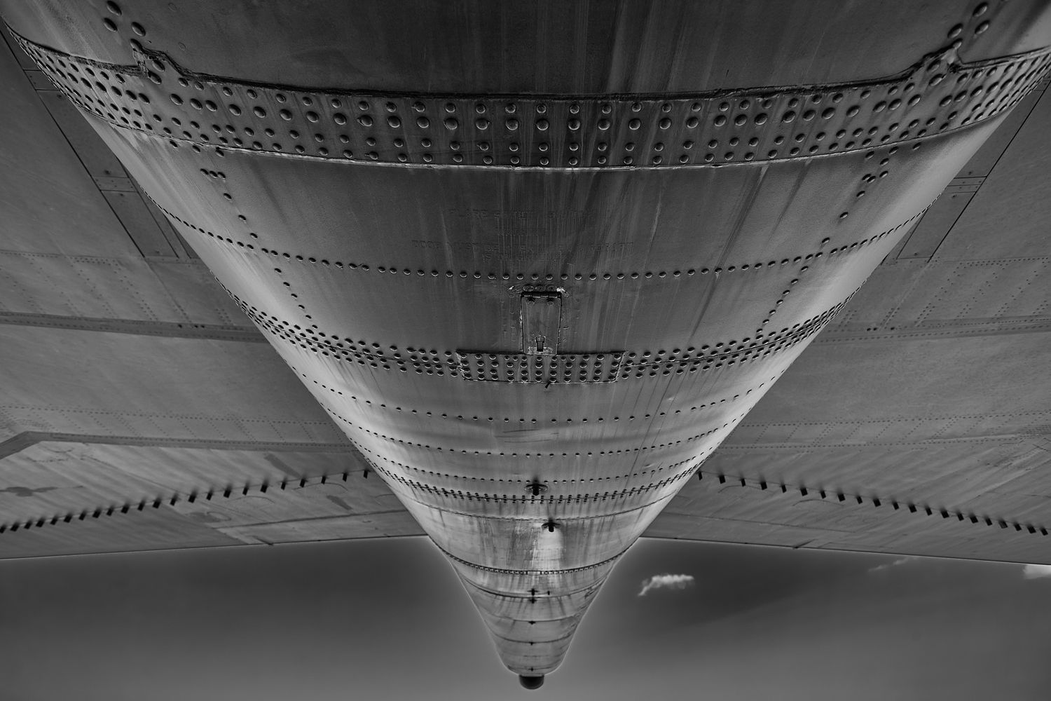 Symmetrical black and white view looking up at a riveted aircraft fuselage tapering toward the tail under a gray sky.