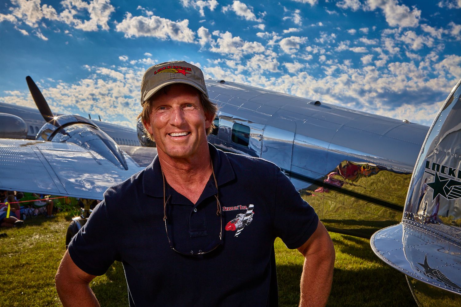 Smiling man in a cap and dark polo shirt stands on a grassy airfield beside a vintage propeller aircraft under a bright blue sky.