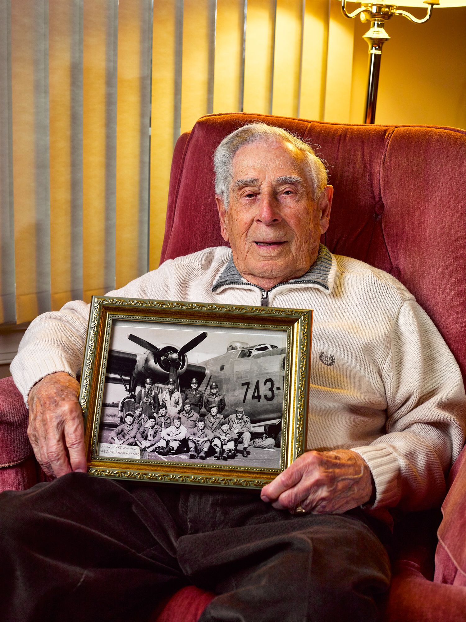 Elderly man in a cream sweater sits in a red armchair holding a framed black-and-white photo of a military aircrew in front of a bomber.