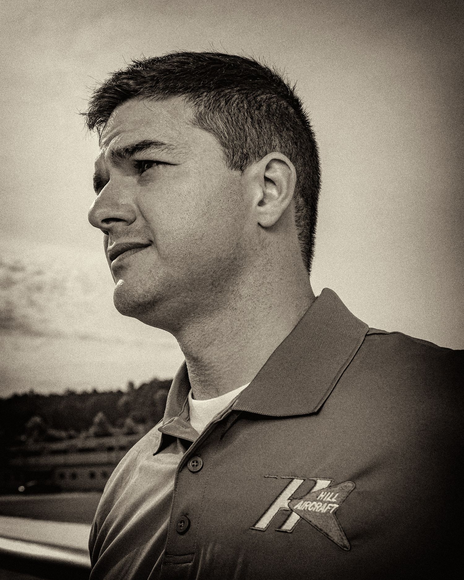 Sepia-toned profile portrait of a man with short dark hair looking into the distance, wearing a collared polo shirt with a logo.