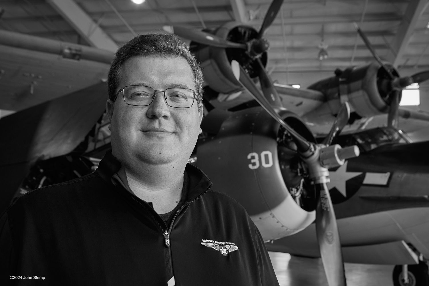 Black and white portrait of a smiling man in glasses standing in a hangar in front of a vintage propeller aircraft numbered 30.