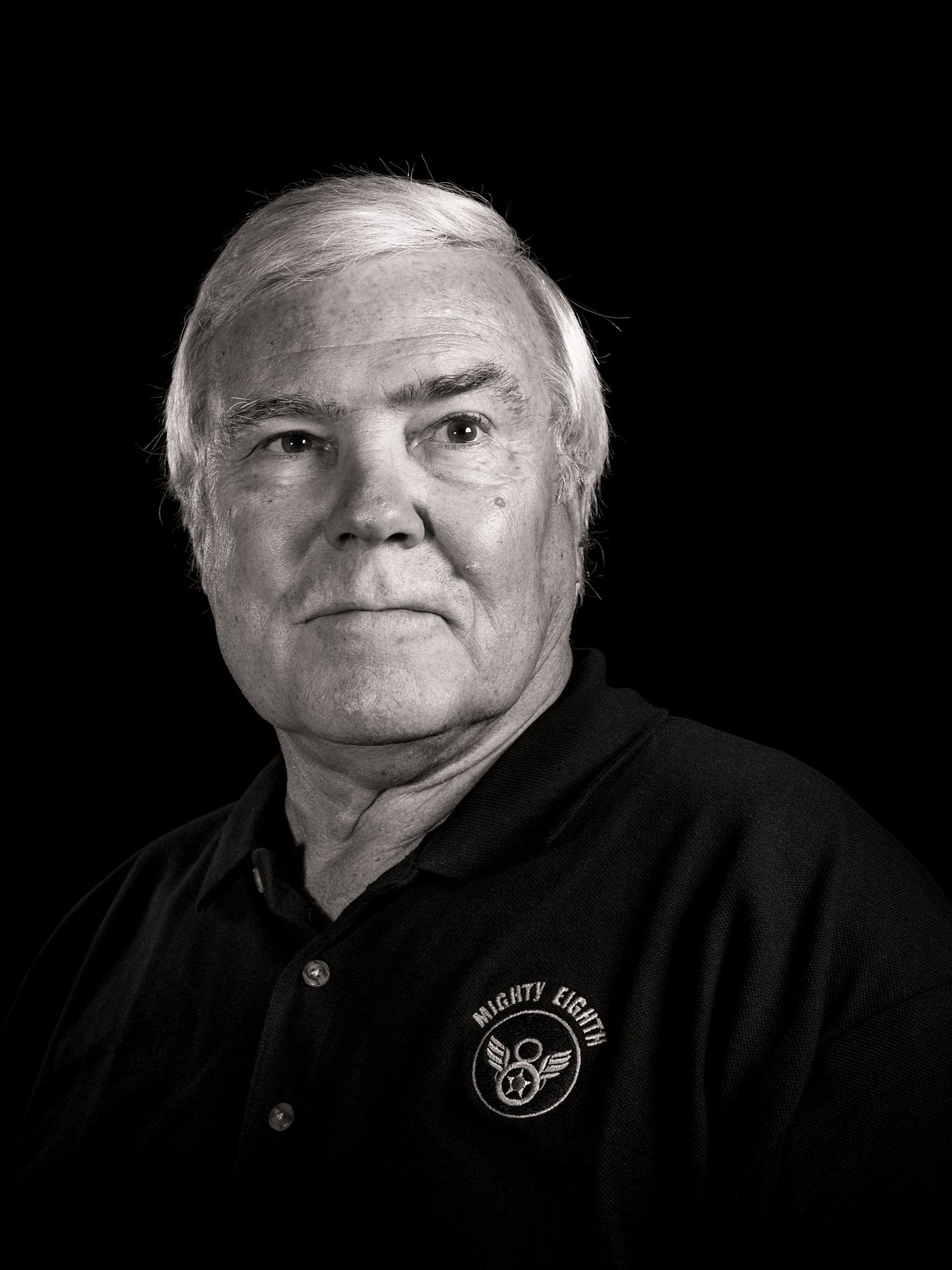 Black and white portrait of a smiling older man with white hair wearing a black polo shirt with a Mighty Eighth logo.
