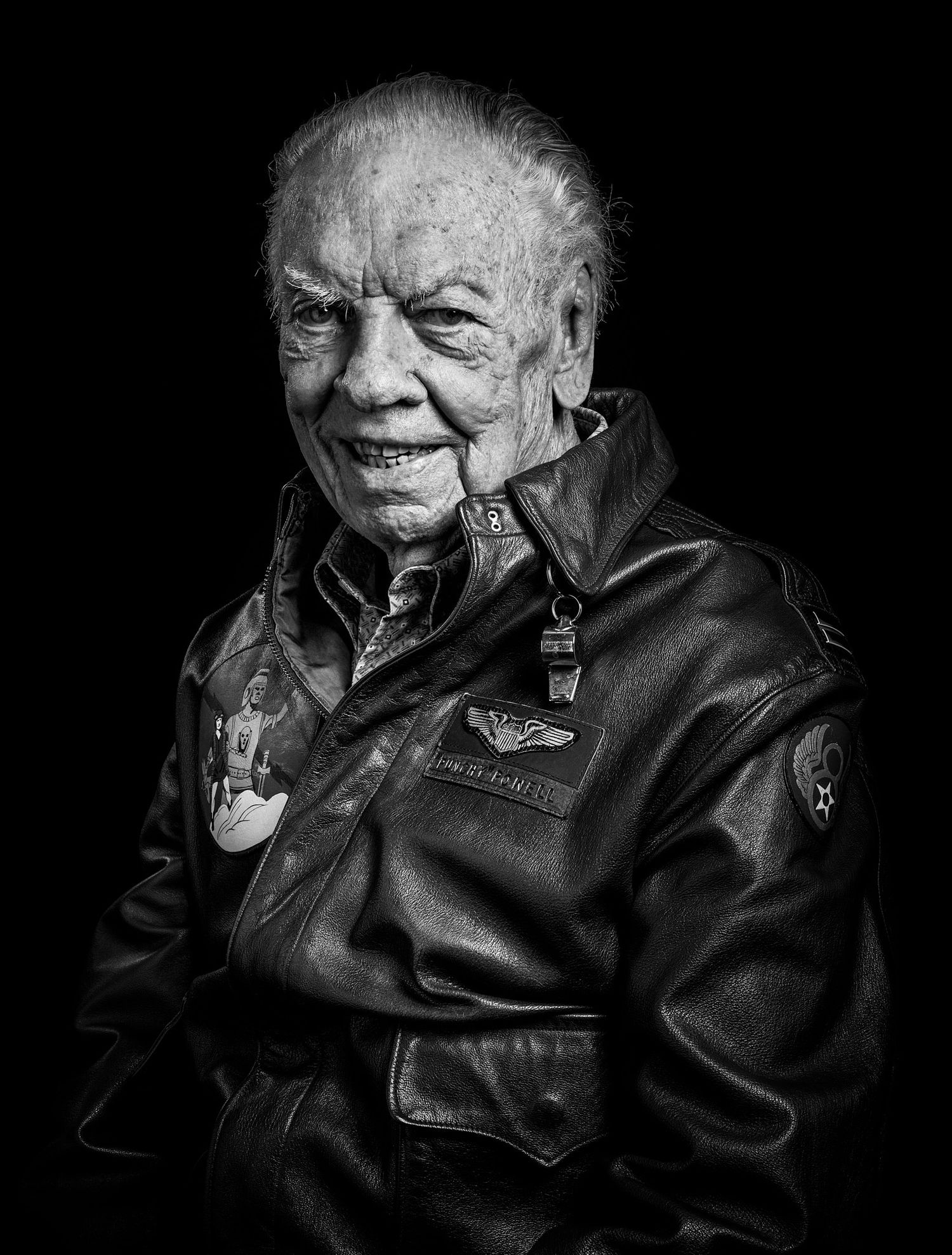 Black and white portrait of a smiling elderly veteran in a worn leather flight jacket with pilot wings and squadron patches.