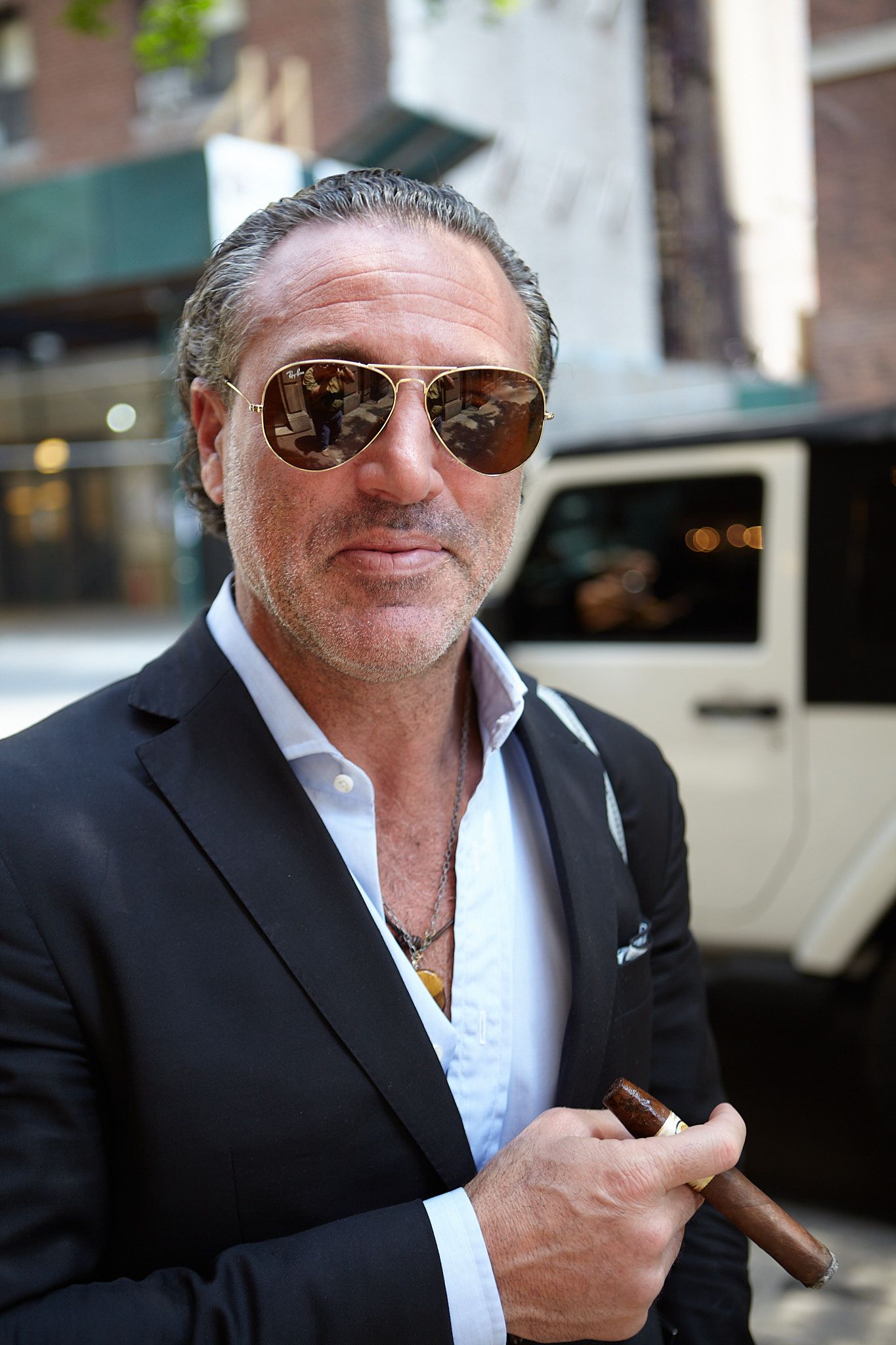 Man with slicked-back gray hair, aviator sunglasses and a navy blazer holds a cigar on a city street beside a white vehicle.