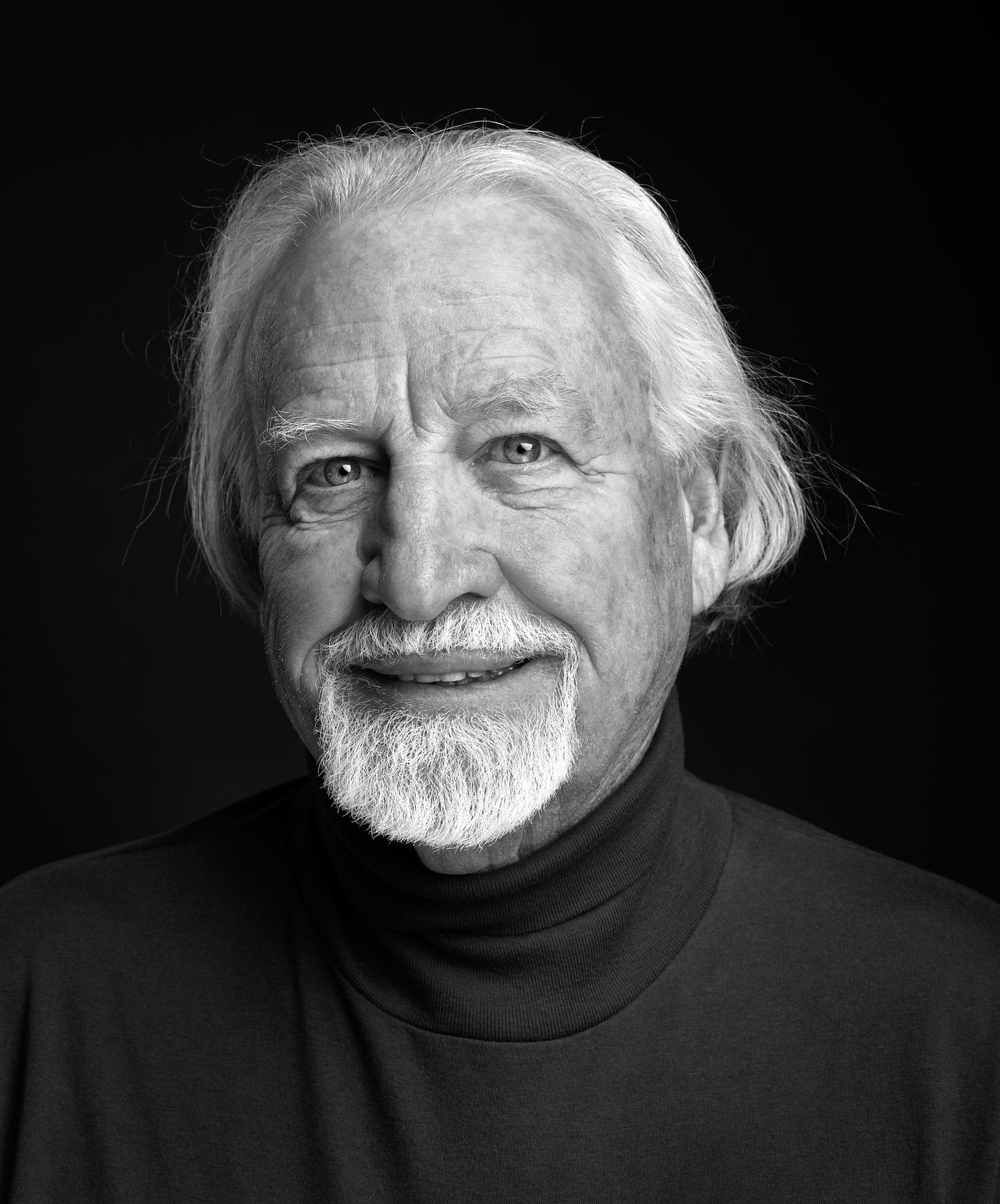 Black and white portrait of an older man with swept-back white hair and a goatee, wearing a dark turtleneck sweater.