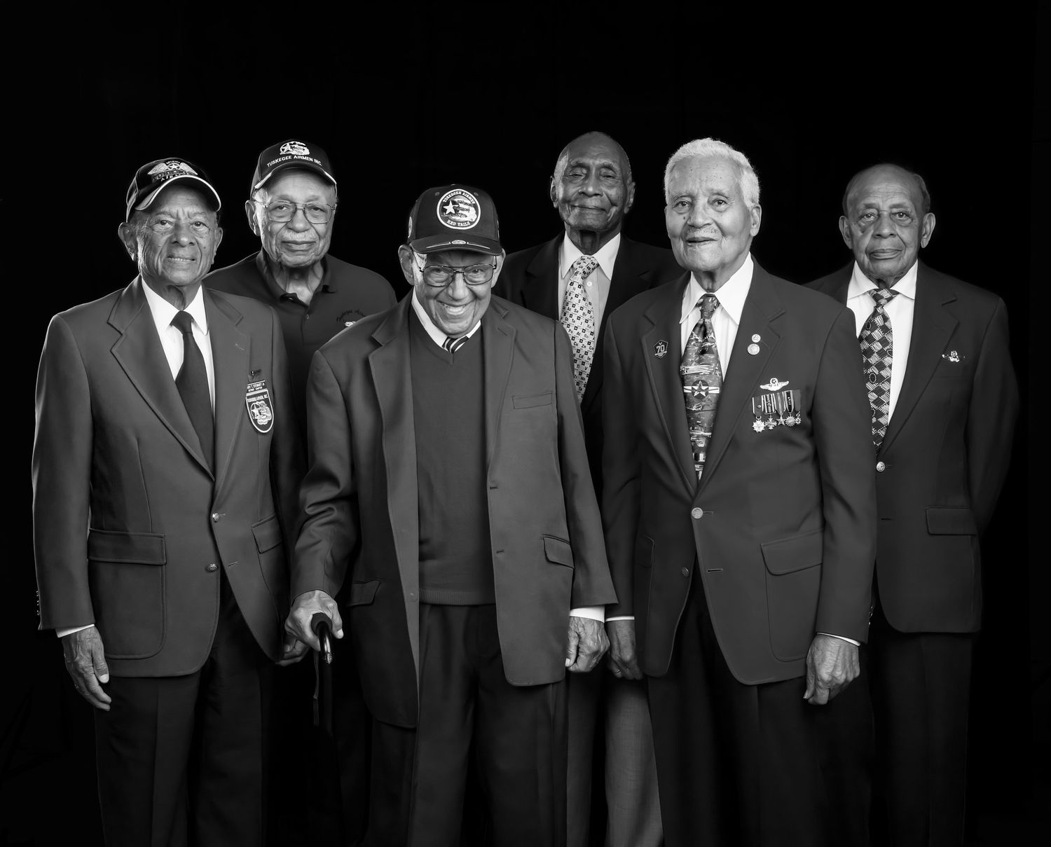 Six elderly men in suits and military caps stand together in a black and white group portrait, some wearing medals and pins.