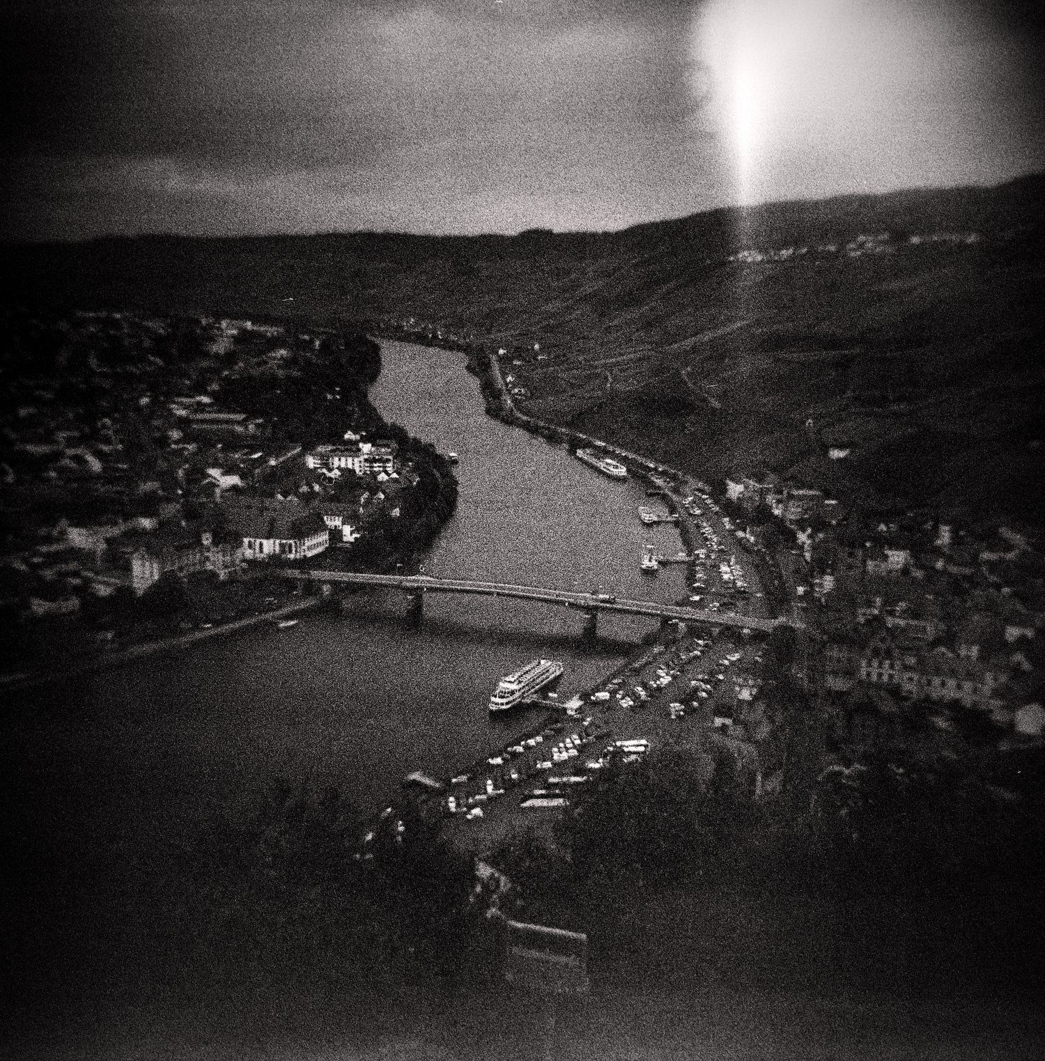 Black and white aerial view of a river winding through a town with a bridge, boats and hillside vineyards under a dramatic sky.