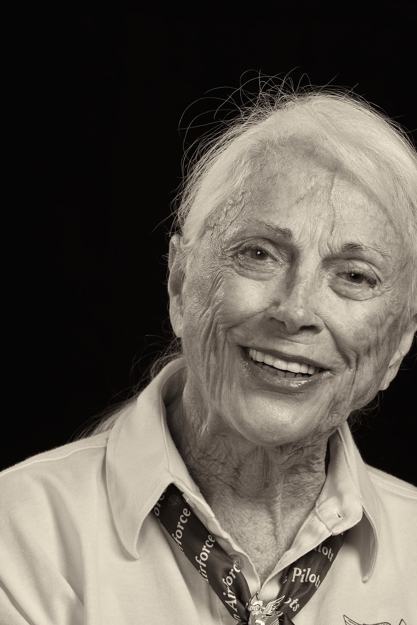 Black and white portrait of a smiling elderly woman with windswept white hair wearing a collared shirt and an Air Force pilot scarf.