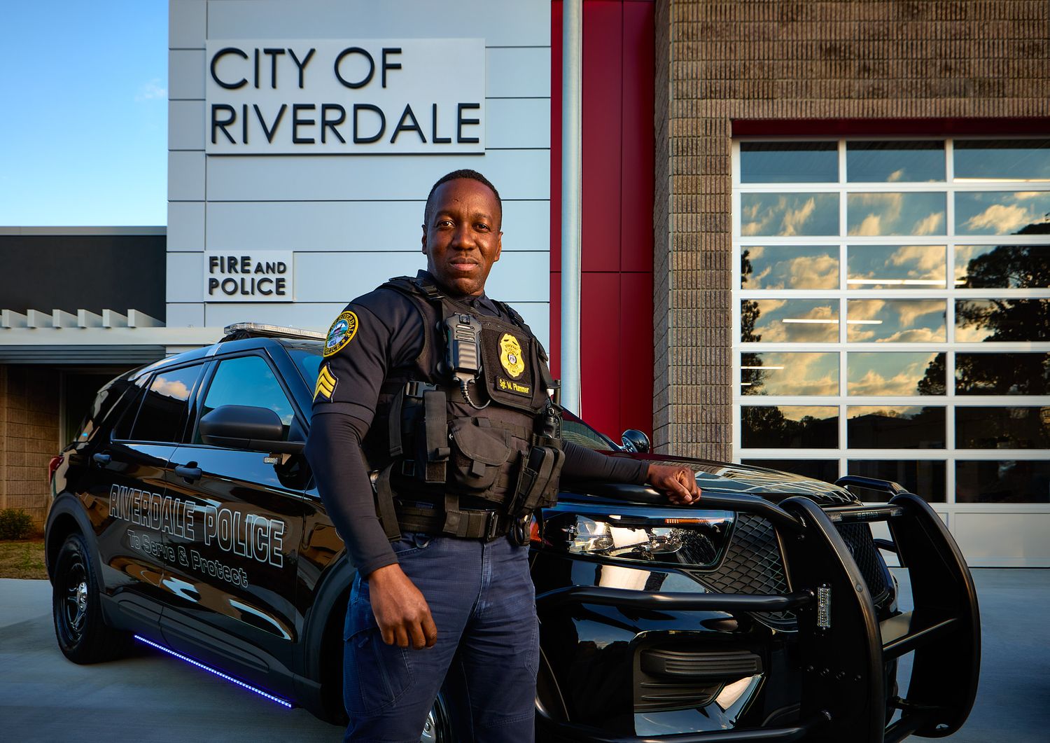 Police officer in uniform and tactical vest stands beside a black patrol SUV outside the City of Riverdale Fire and Police building.