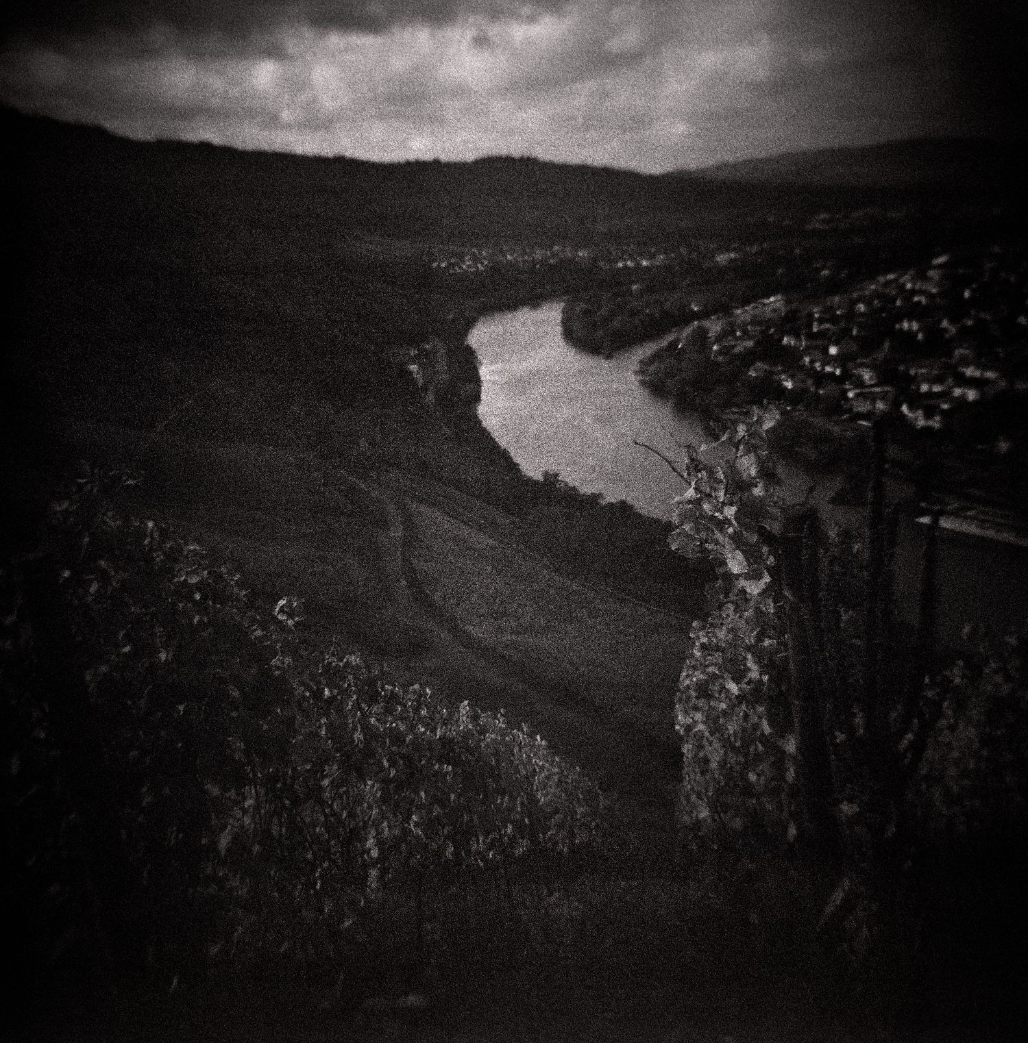 Moody black and white landscape of a winding river curving through a valley with vineyards, hillside village, and dramatic cloudy skies.
