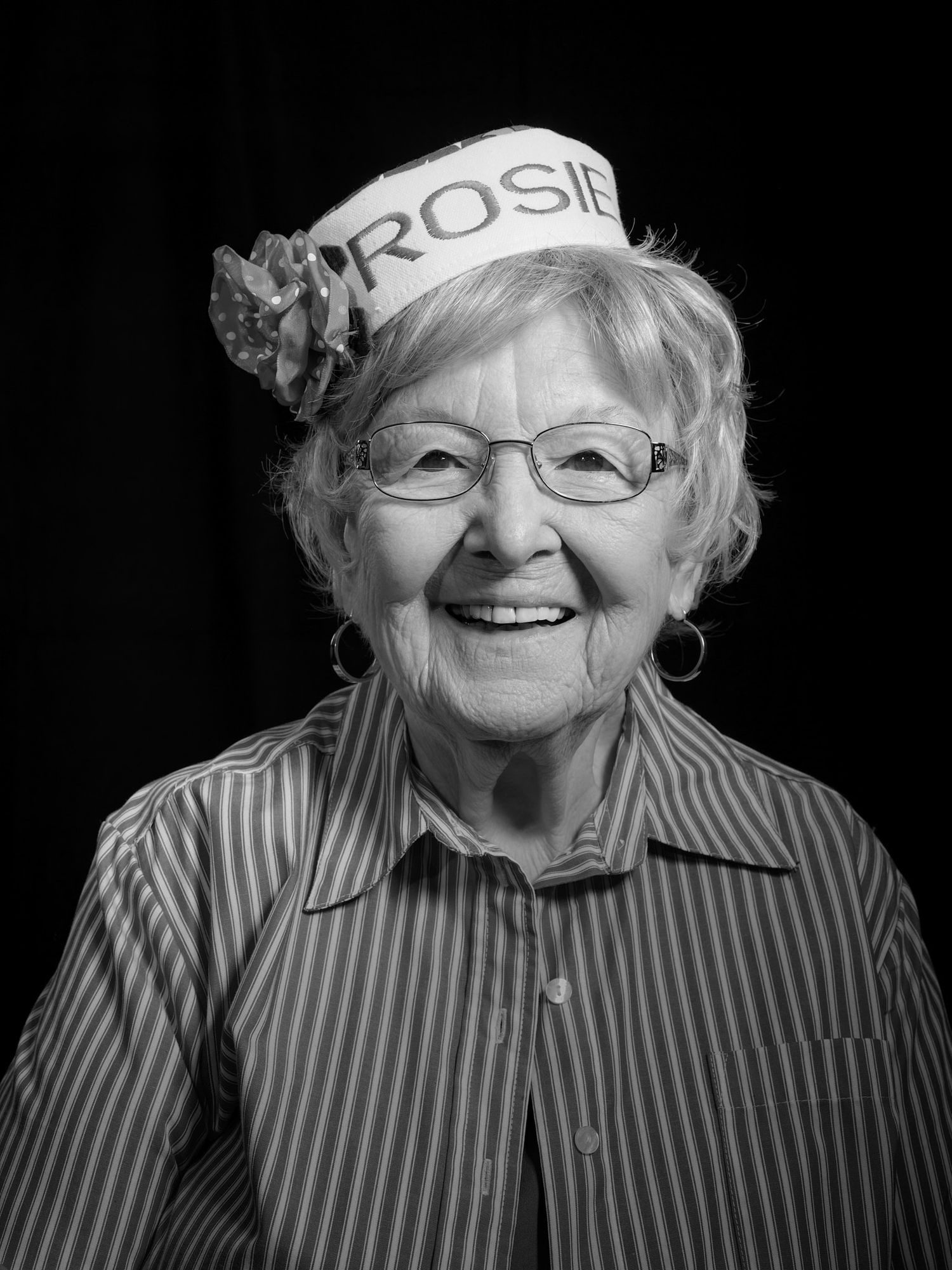 Black and white portrait of a smiling elderly woman with glasses wearing a decorated Rosie the Riveter sailor-style hat.