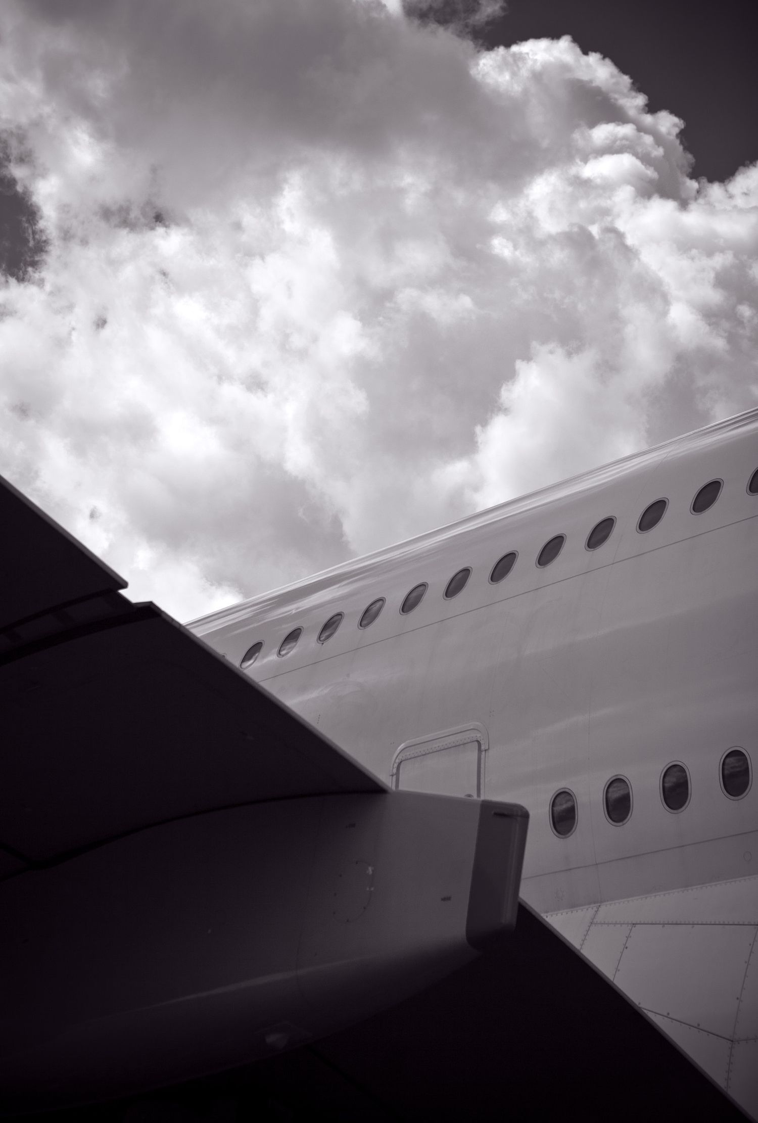 Black and white photo of a large airplane fuselage with a row of passenger windows beneath a dramatic cloudy sky.