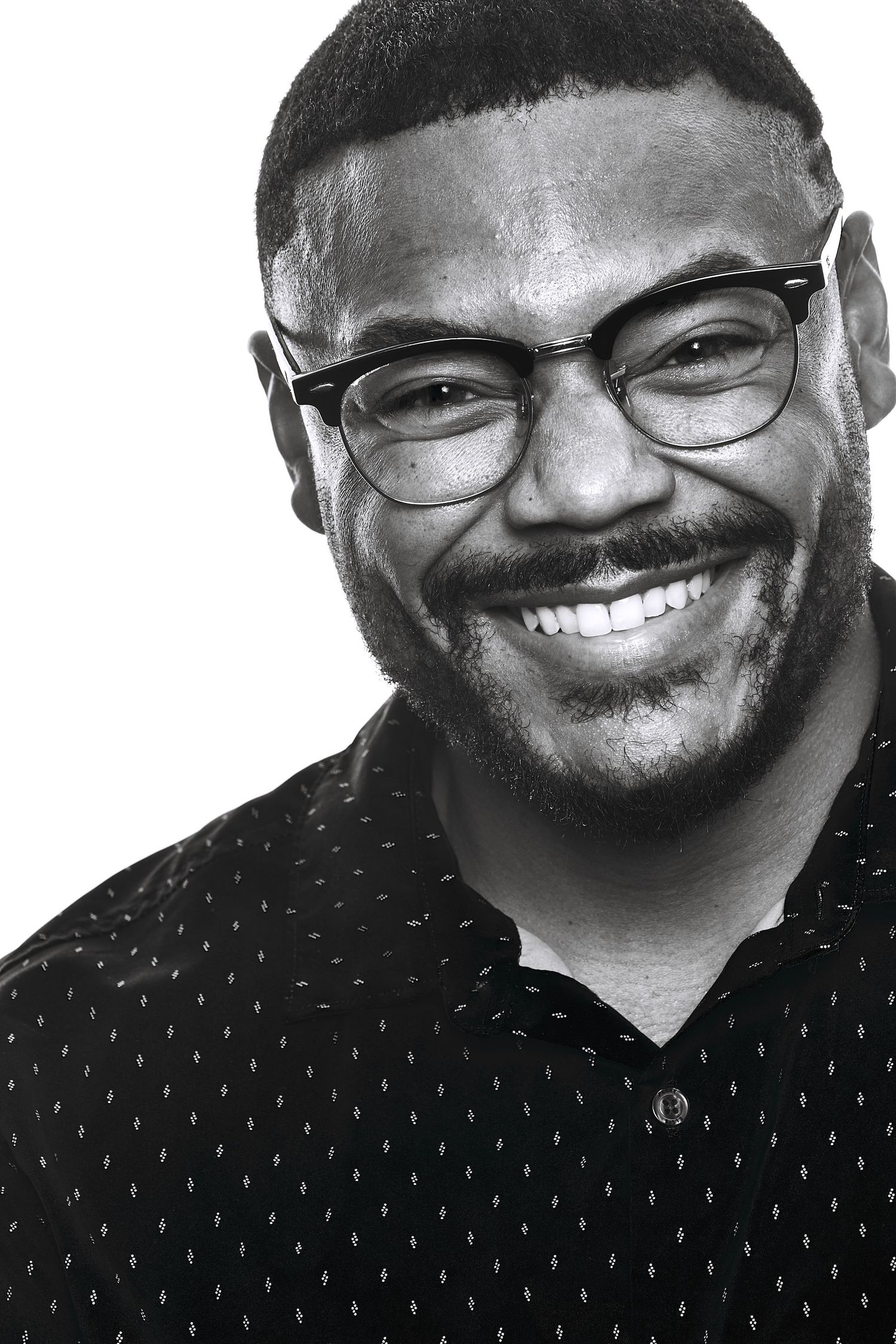 Black and white portrait of a smiling bearded man in black browline glasses and a dark patterned button-up shirt.