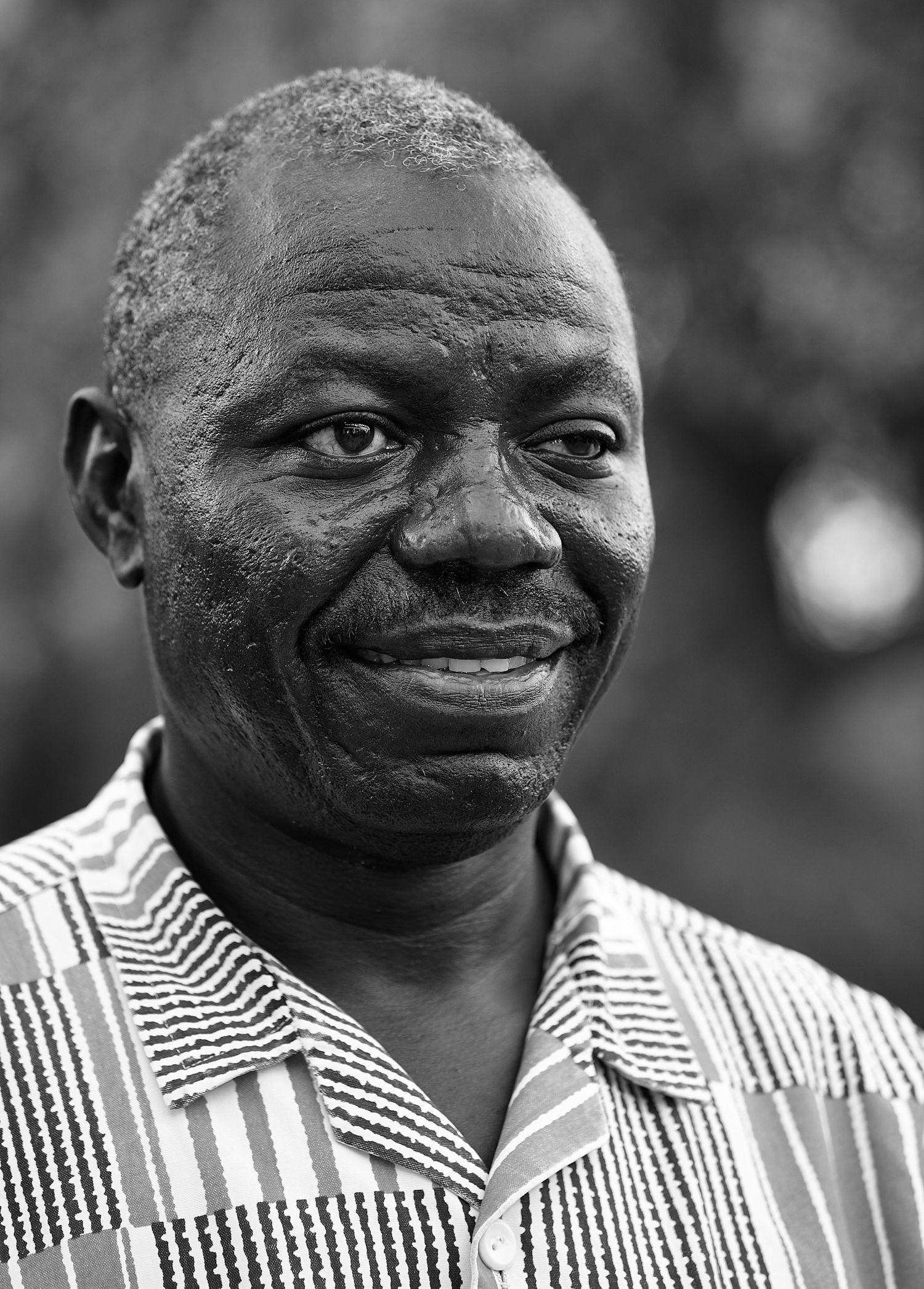 Black-and-white portrait of a smiling middle-aged man with short hair wearing a striped patterned collared shirt outdoors.