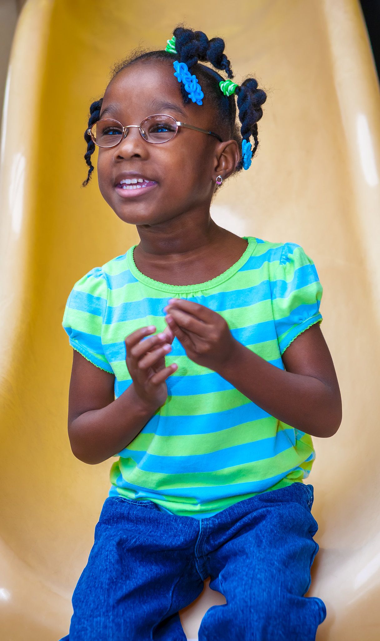 Smiling young girl with glasses and blue and green hair ties wears a striped green top and jeans while sitting on a yellow slide.