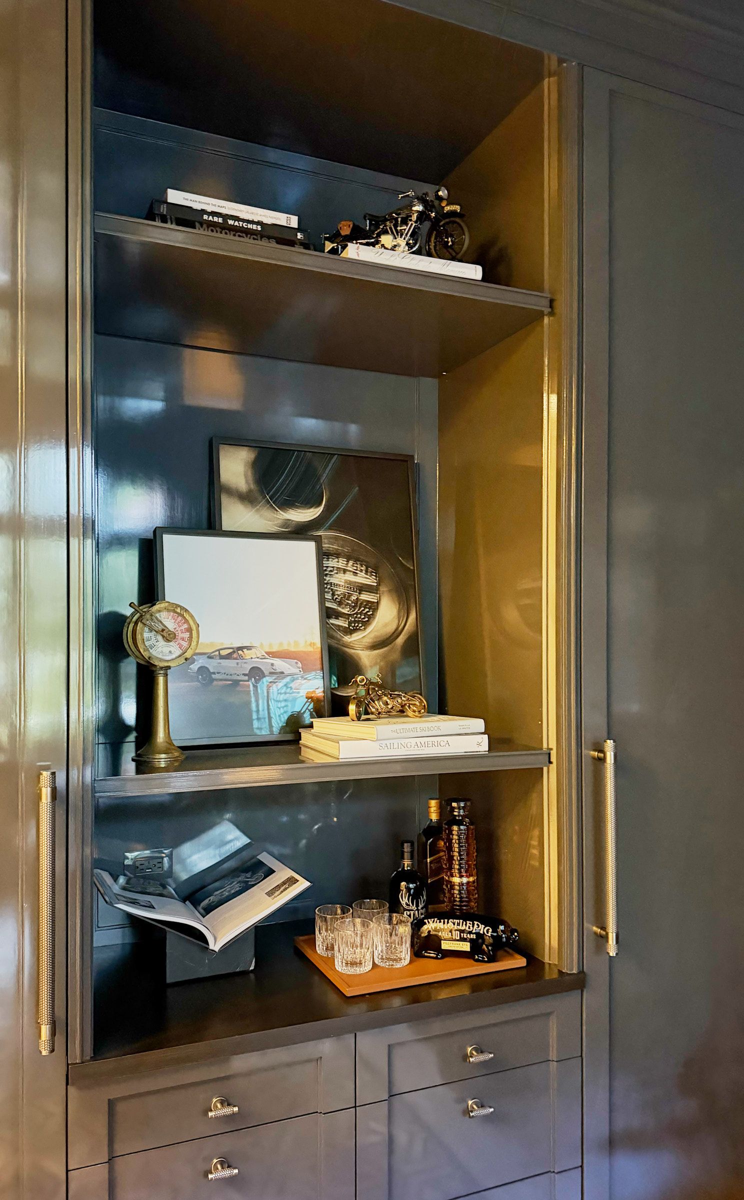 Glossy gray built-in cabinet with brass handles, styled shelves holding books, framed art, a vintage clock and a small bar tray.