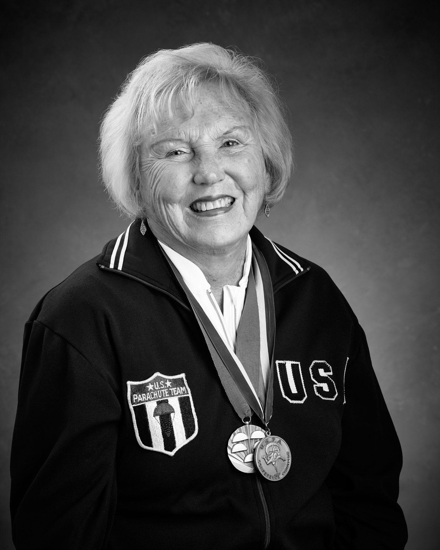 Smiling older woman in a black USA U.S. Parachute Team jacket wearing two medals around her neck in a black-and-white studio portrait.
