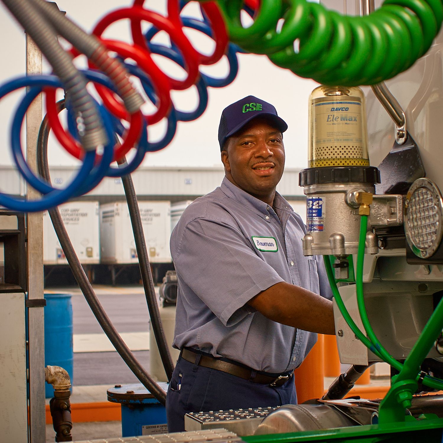 Smiling mechanic in a blue cap and gray uniform works at a service bay surrounded by coiled red, blue and green air hoses and equipment.