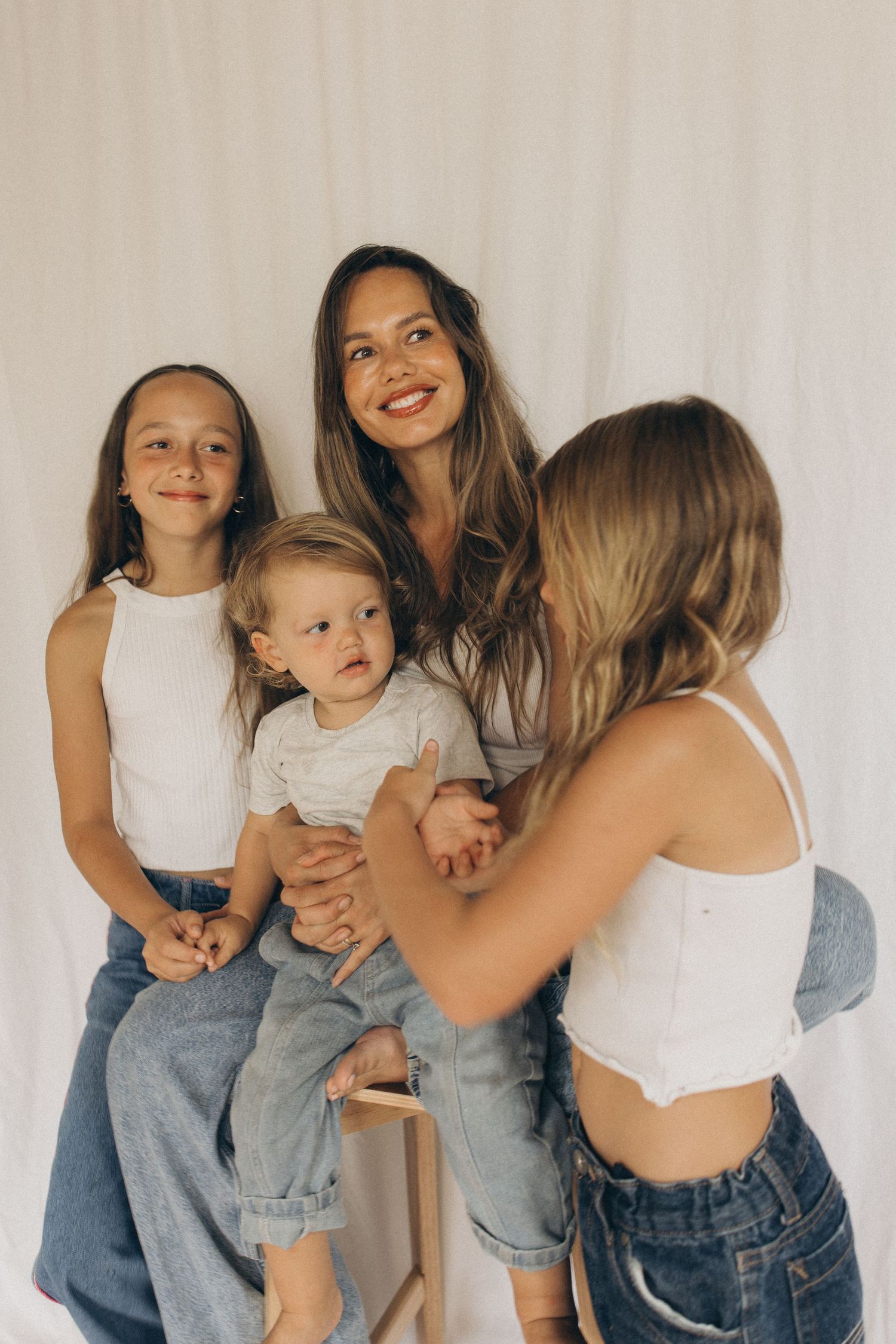 Mother smiles with three children including toddler and two daughters in white tops against white curtain backdrop in New Zealand.