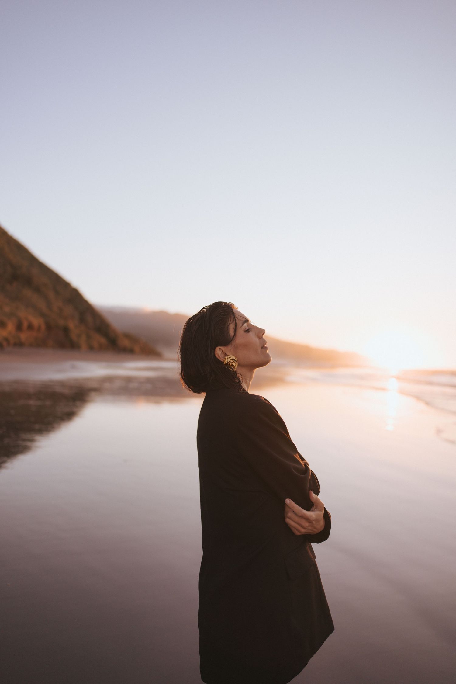 Woman in black blazer with closed eyes embracing herself on a serene reflective beach at sunset in New Zealand.