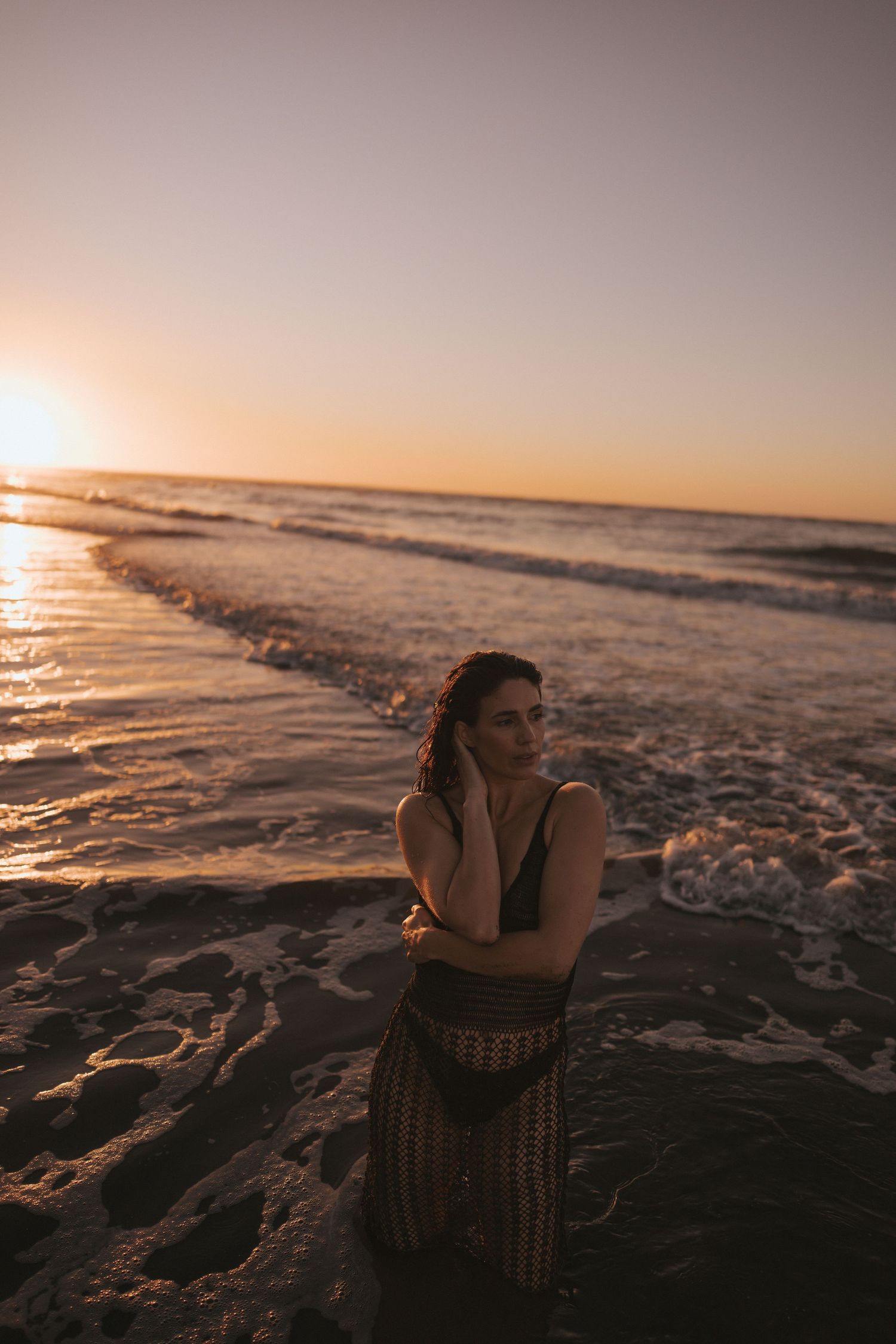 Woman in crochet dress standing in ocean waves on sandy beach during vibrant golden sunset in New Zealand.