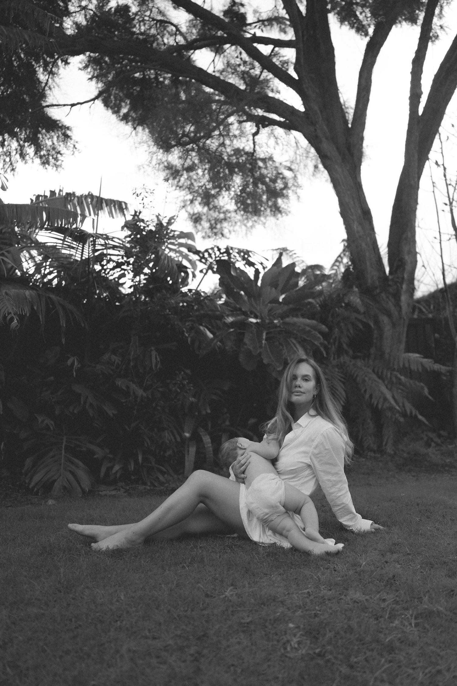 Black and white photo of woman sitting on grass holding child under large tree in New Zealand.